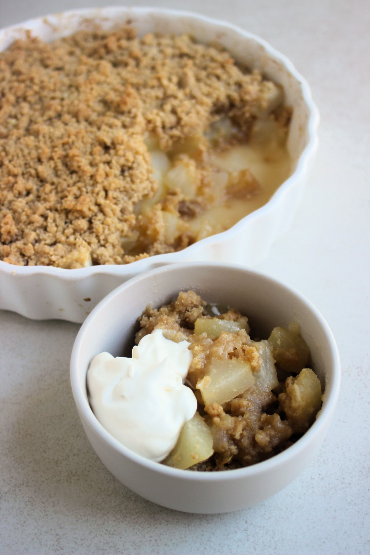 Small bowl with pear crumble and whipped cream, and a round baking dish with pear crumble behind.
