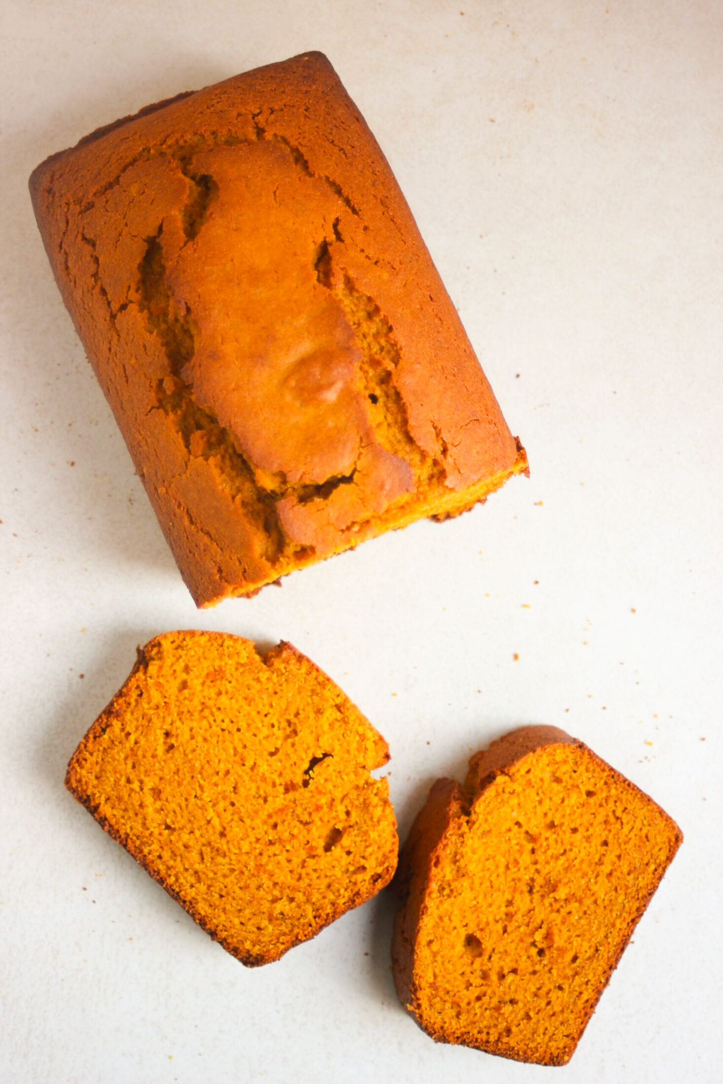 A pumpkin bread and two slices of it on a white surface seen from above.