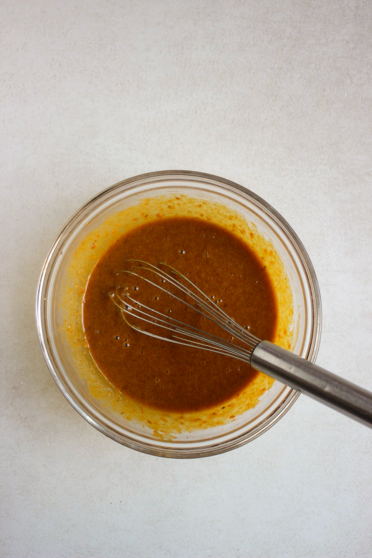 Glass bowl with a brown liquid mixture and a hand whisk.