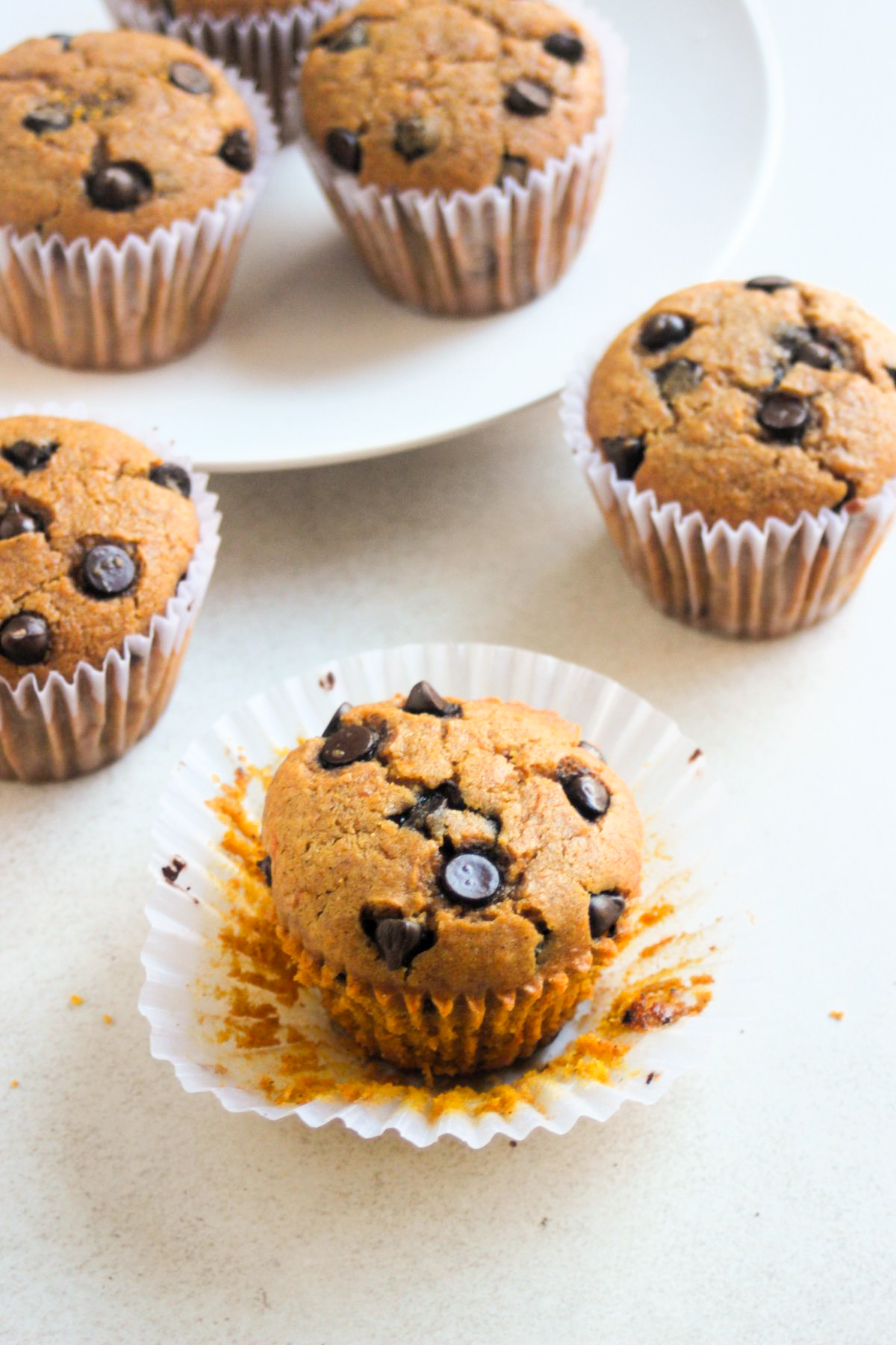 Pumpkin muffins with chocolate chips, with the liner open on a white surface. More muffins, behind and in a white plate.