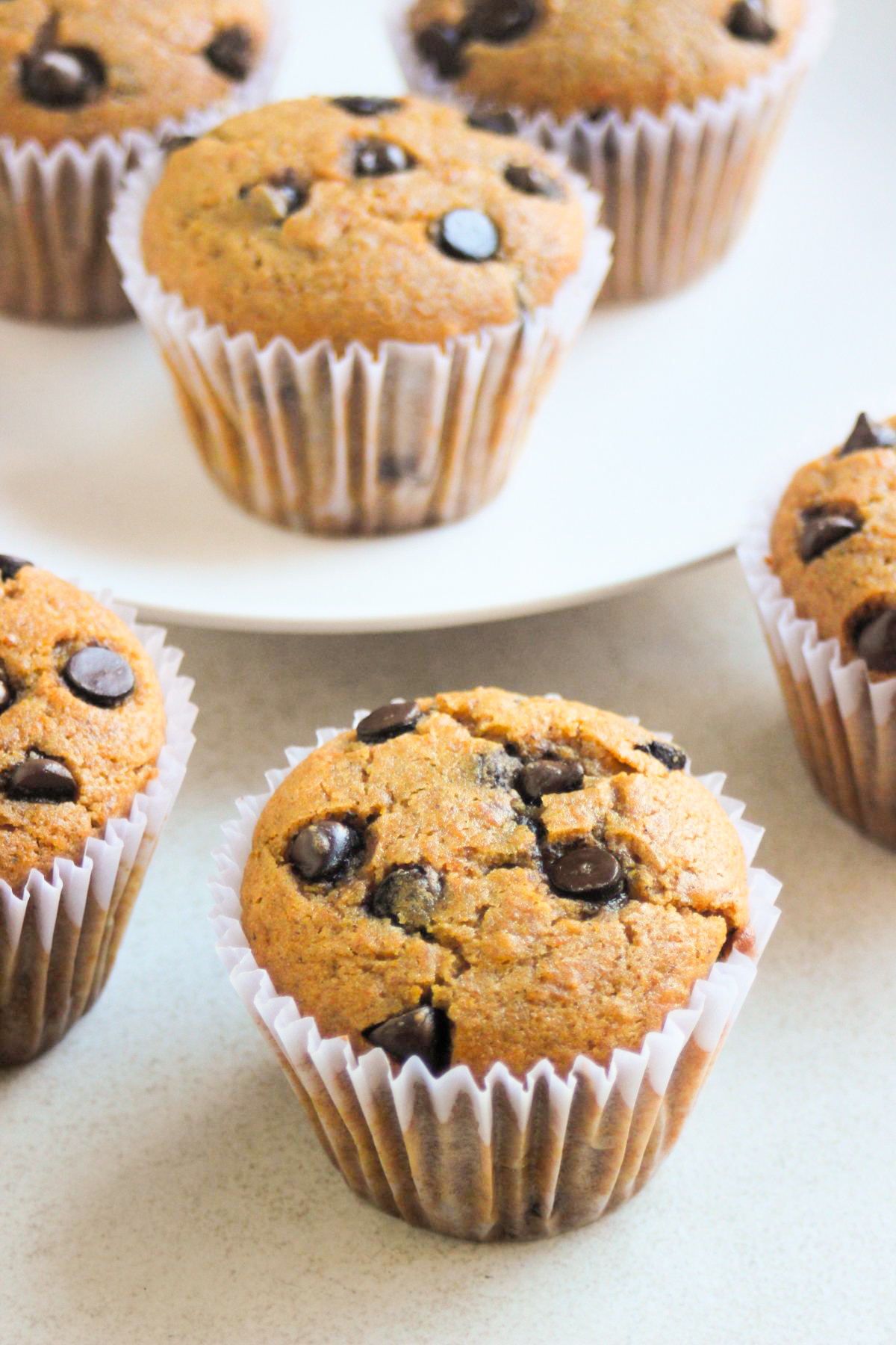 Pumpkin muffins with chocolate chips and liners on a white surface. More muffins are behind on a white plate.