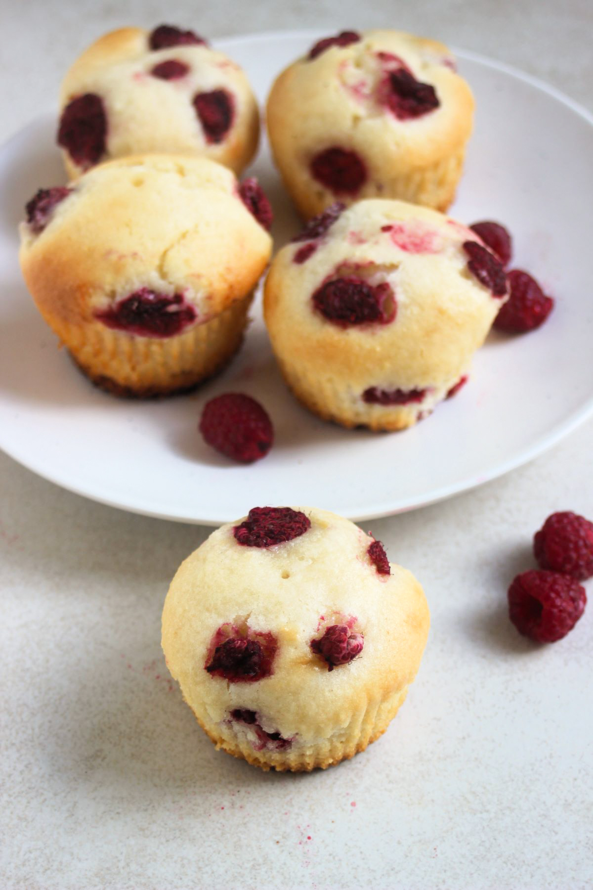 Raspberry muffin and fresh raspberries on a white surface. More muffins behind on a white plate and fresh raspberries.