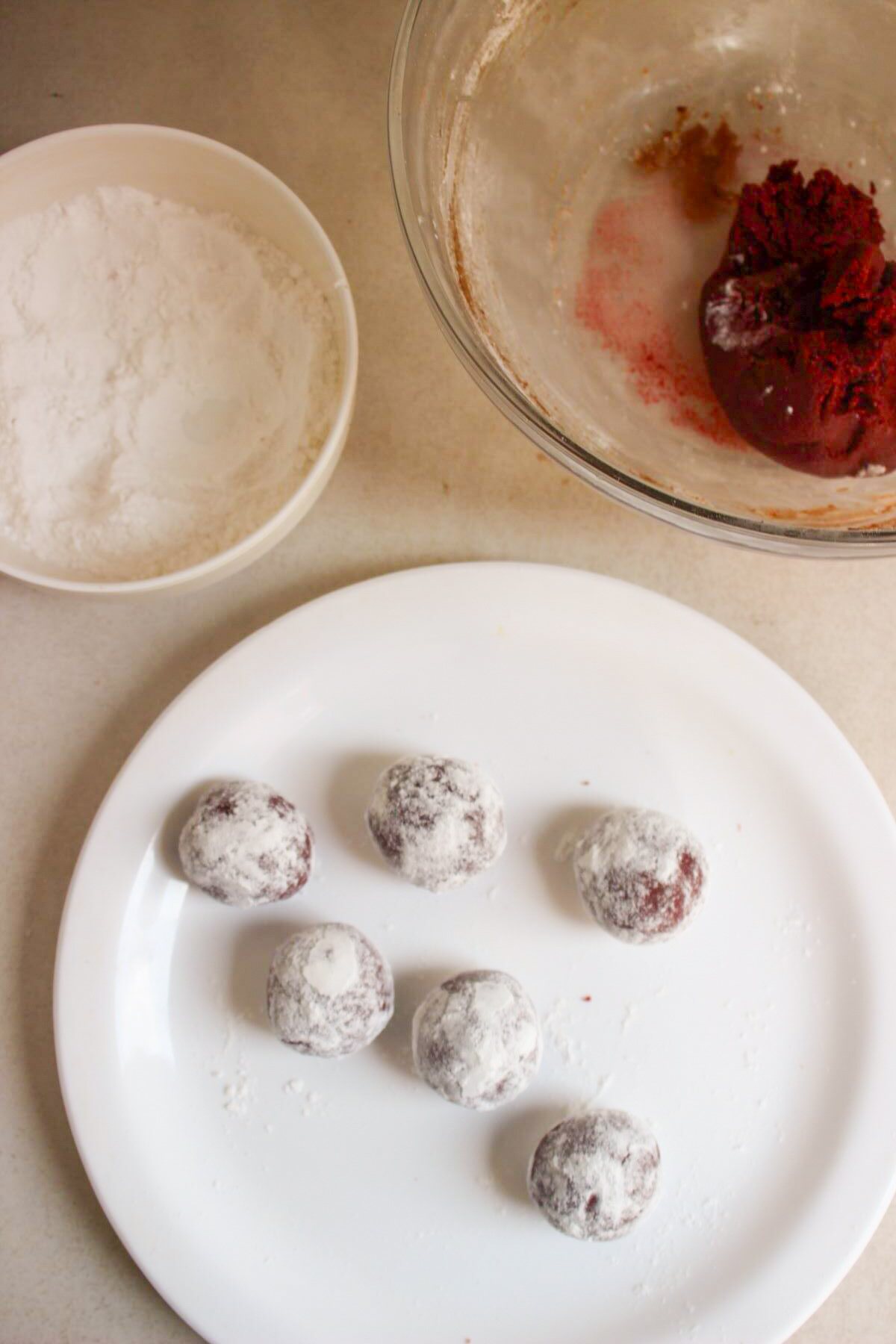 Plate with cookie dough balls coated with powdered sugar. Besides, a small bowl with powdered sugar and a large bowl with cookie dough.