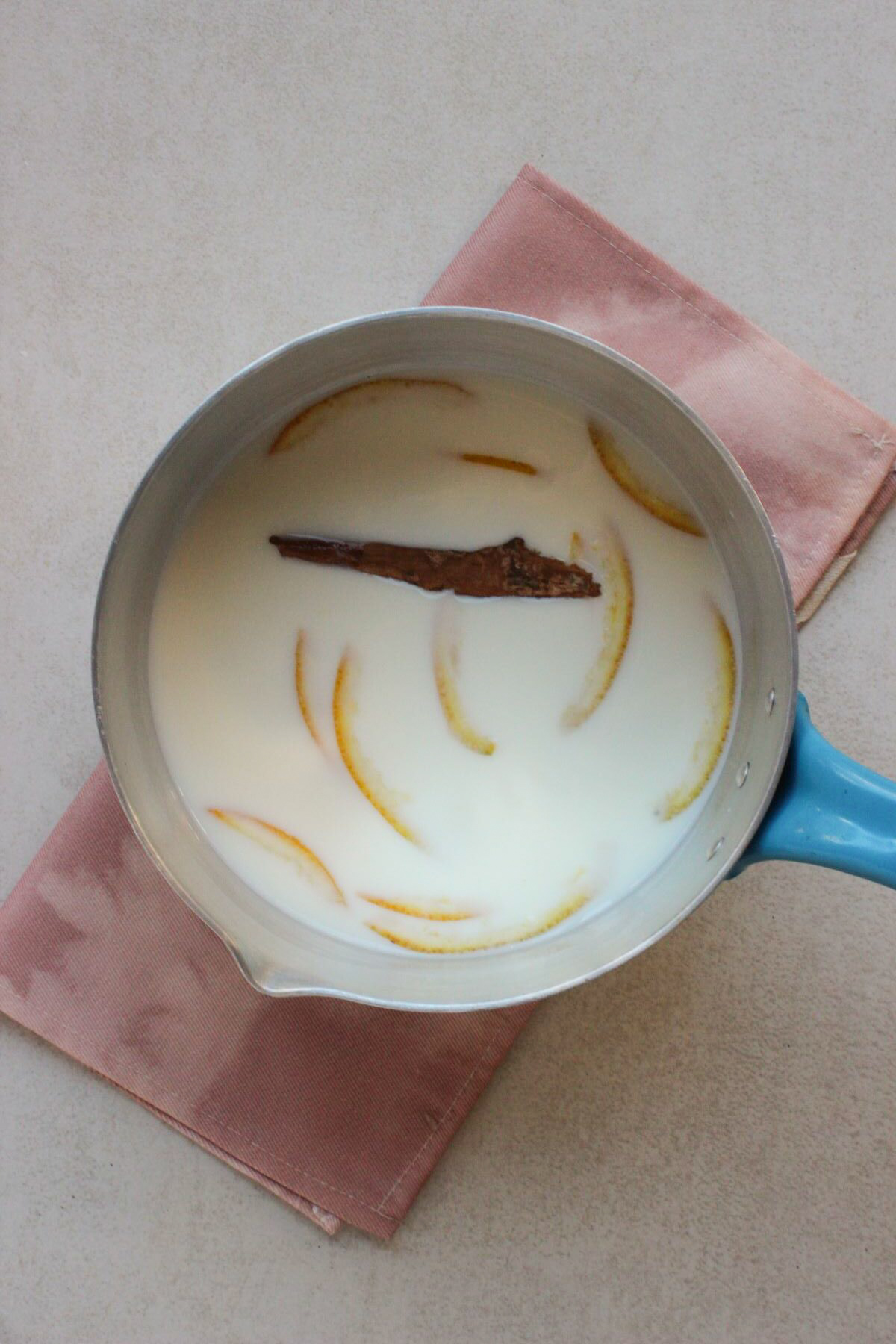 Deep saucepan with a white liquid, cinnamon stick, and orange peels. Pink napkin under the saucepan.