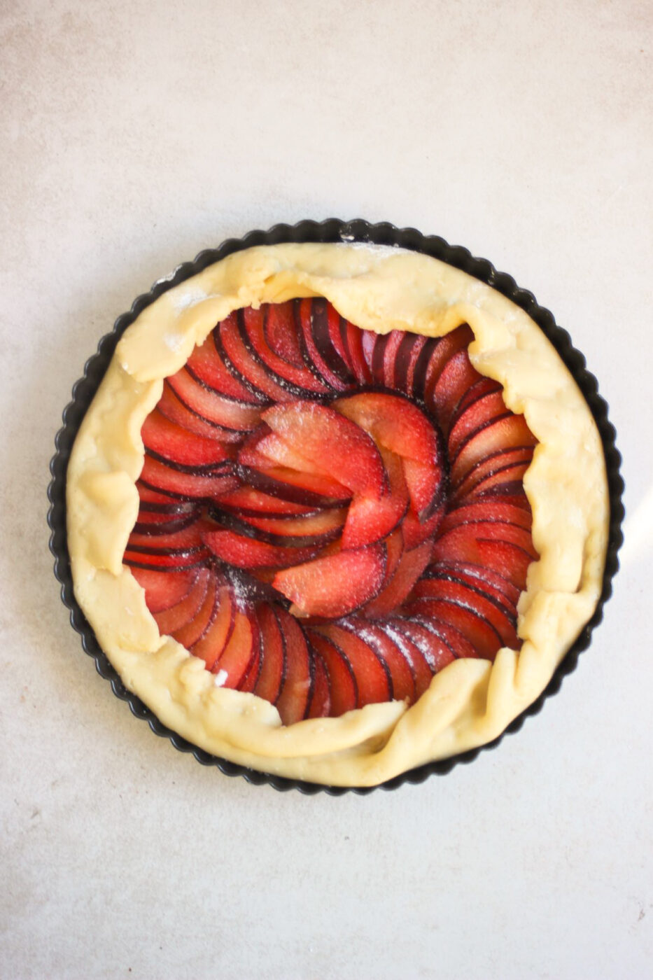 Raw plum tart in a tart pan on a white surface seen from above.