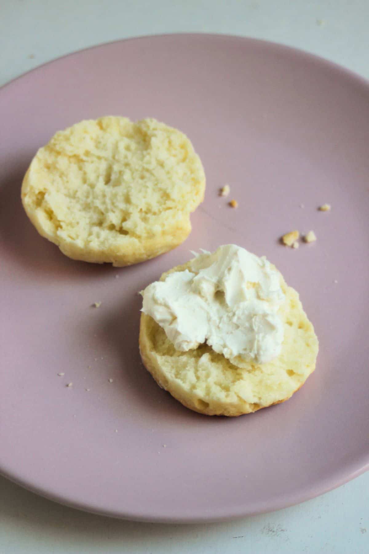 A scone split into two halves on a pink plate. One biscuit is topped with cream cheese.