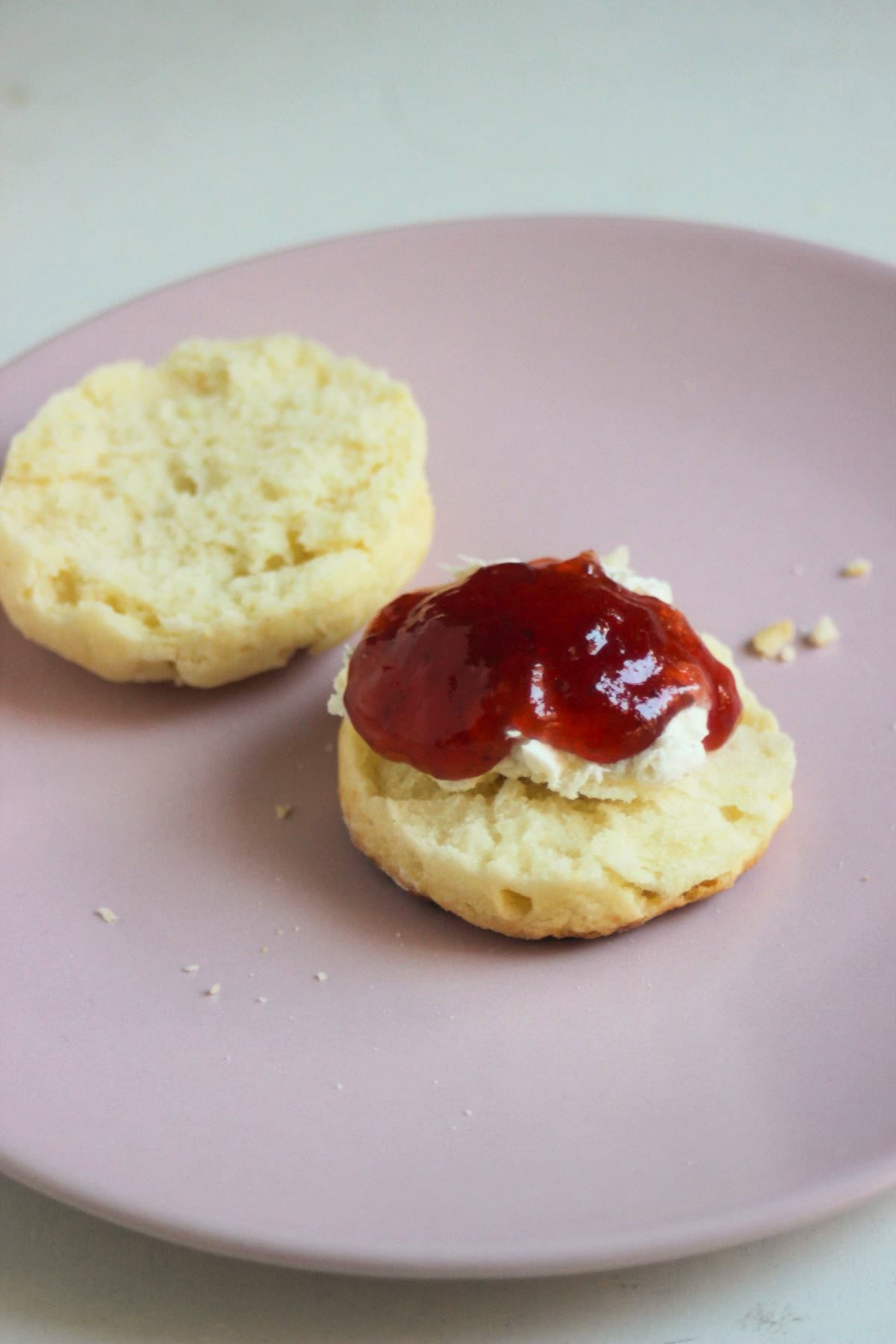 Scon cut in two half on a pink plate. One is topped with cream cheese and jam