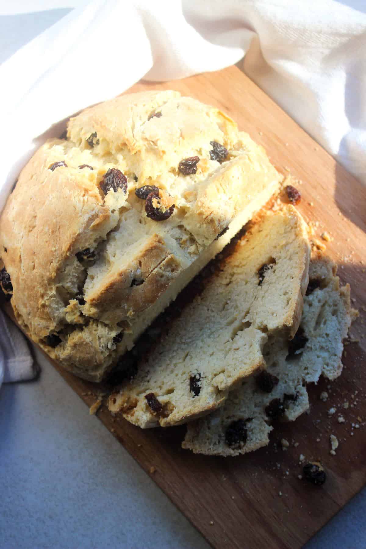 Soda bread with raisins and two slices of it on a wooden board seen from above. White kitchen towel behind.