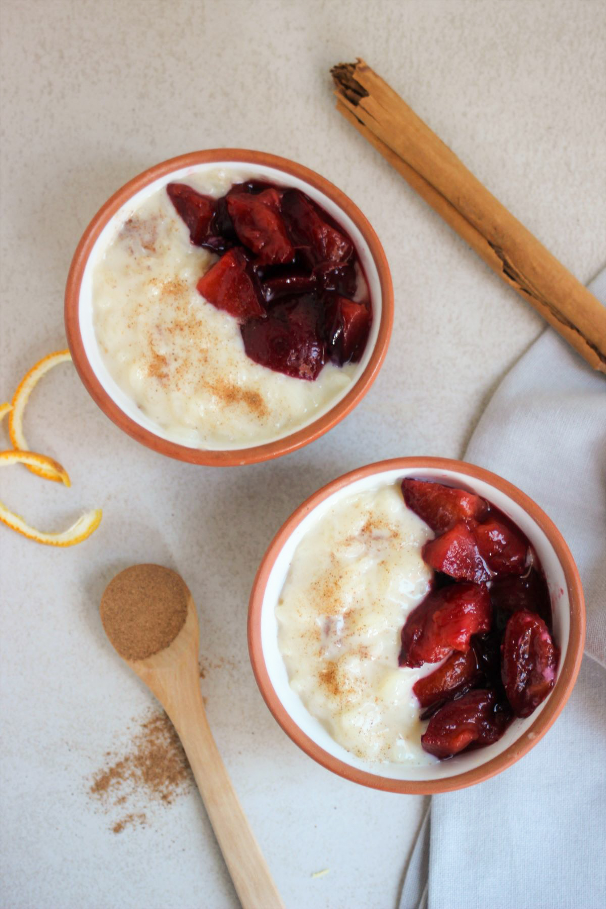 Two tiny bowls with rice pudding and plum compote seen from above. A tablespoon and a cinnamon stick.