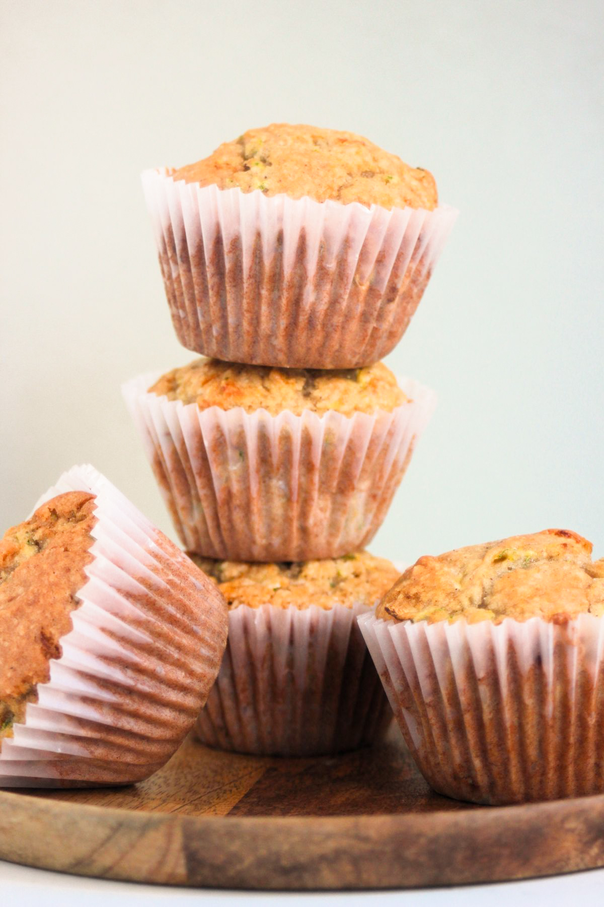 Stack of zucchini muffins with liners on a wooden plate. Two muffins besides. Light grey background.