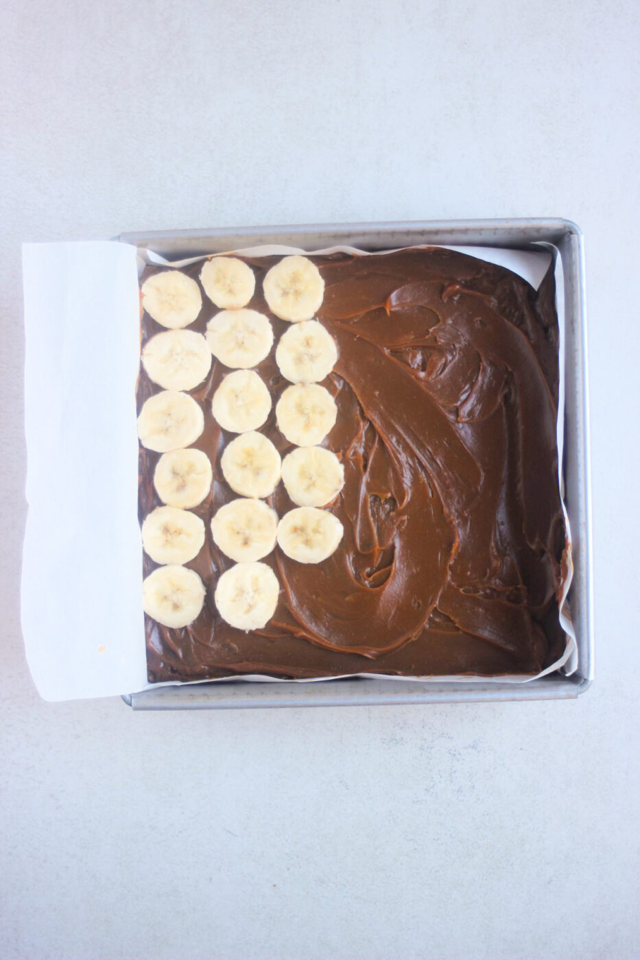 Top view of a square baking pan with parchment paper, dulce de leche, and banana slices arranged on one side.
