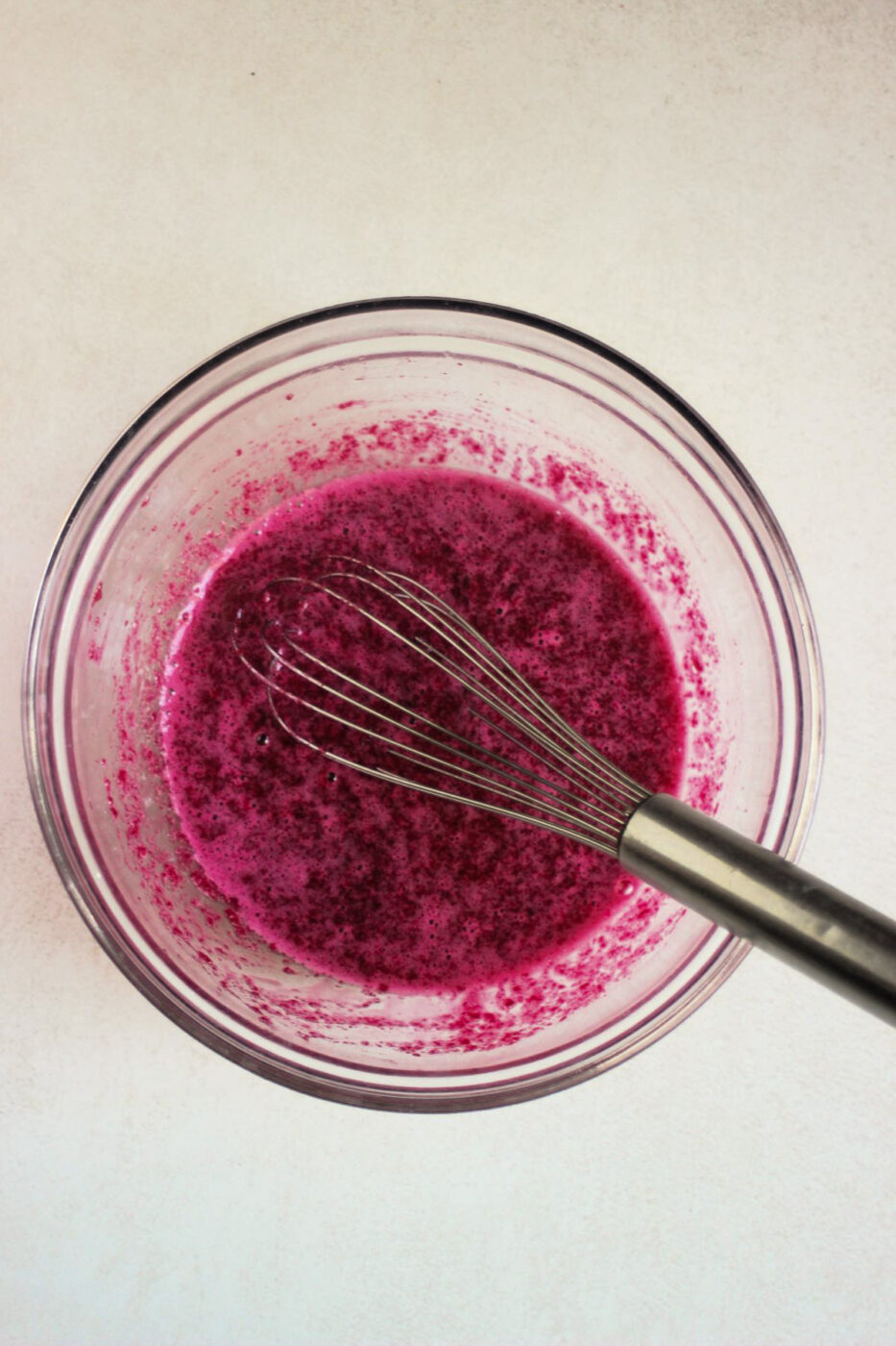 Glass bowl with a magenta colour mixture and a hand whisk.