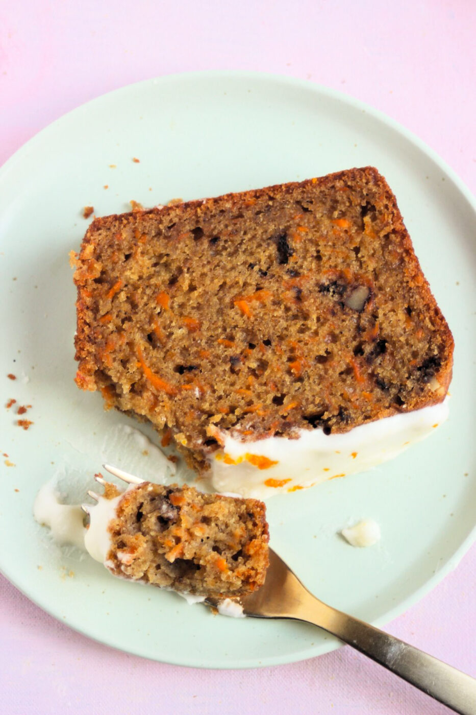 A slice of carrot loaf cake with cream cheese frosting on an aqua-green plate. A golden fork with a pice of the slice.