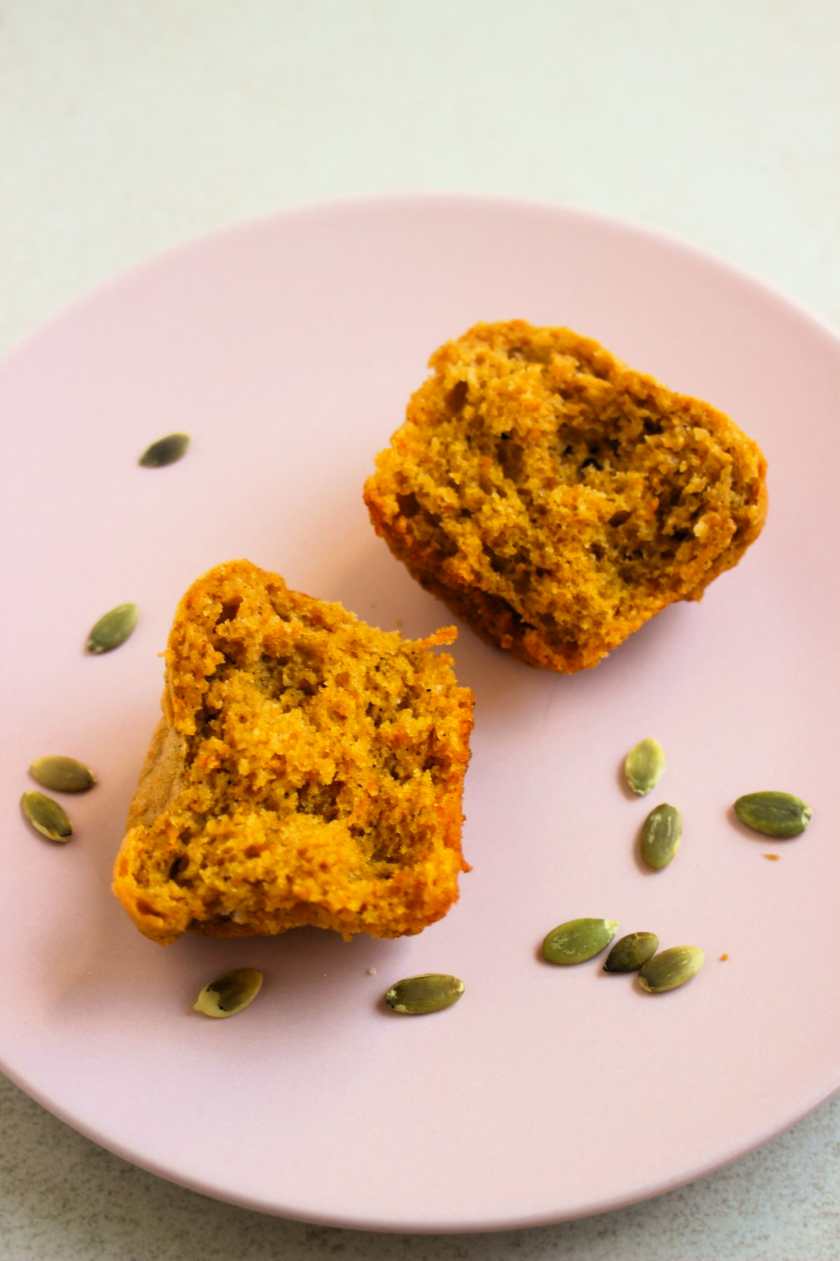 Close-up of a gluten-free pumpkin muffin cut in half on a pink plate. Pumpkin seeds scattered around.