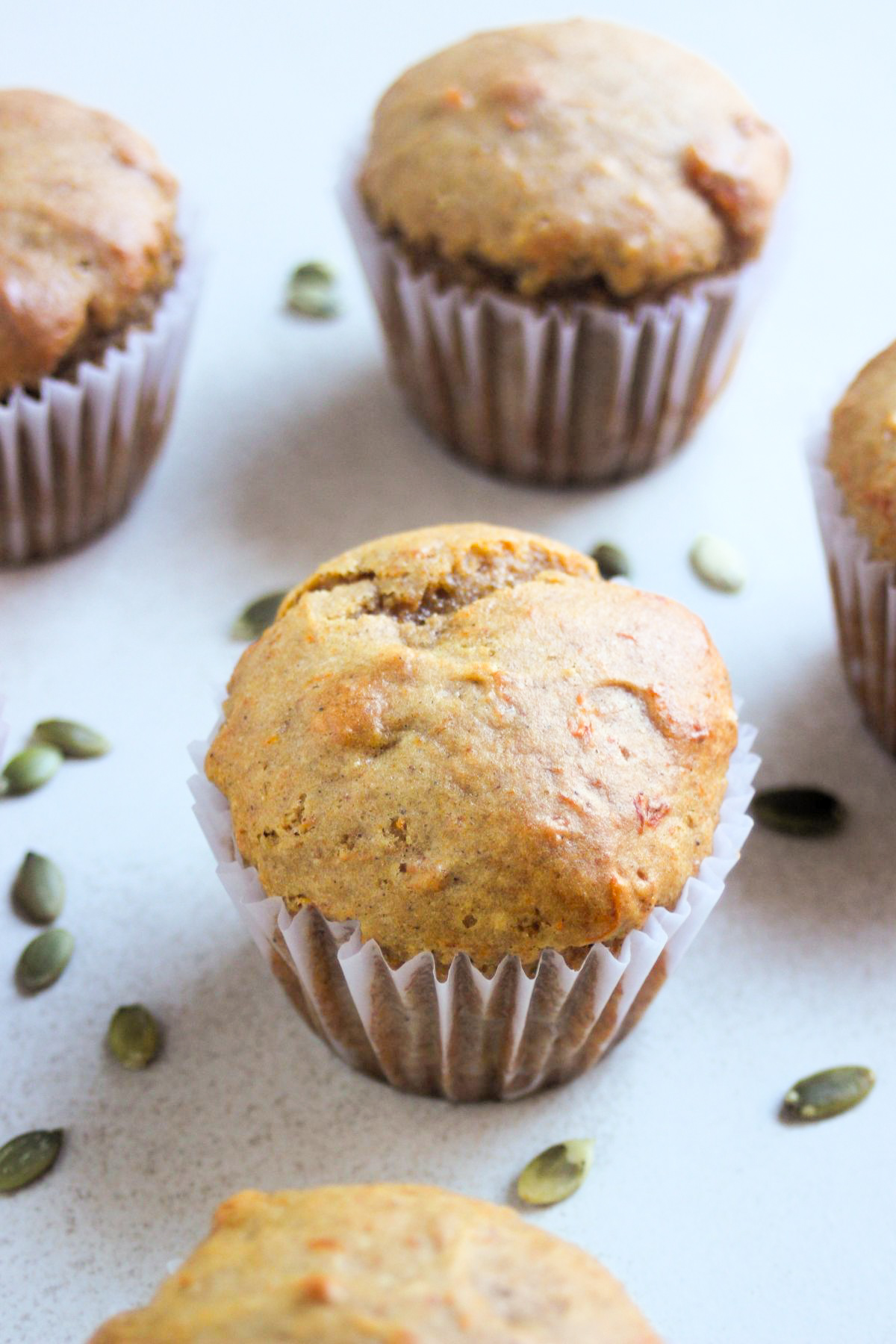 Pumpkin muffins with liners on a white surface and pumpkin seeds spread around.