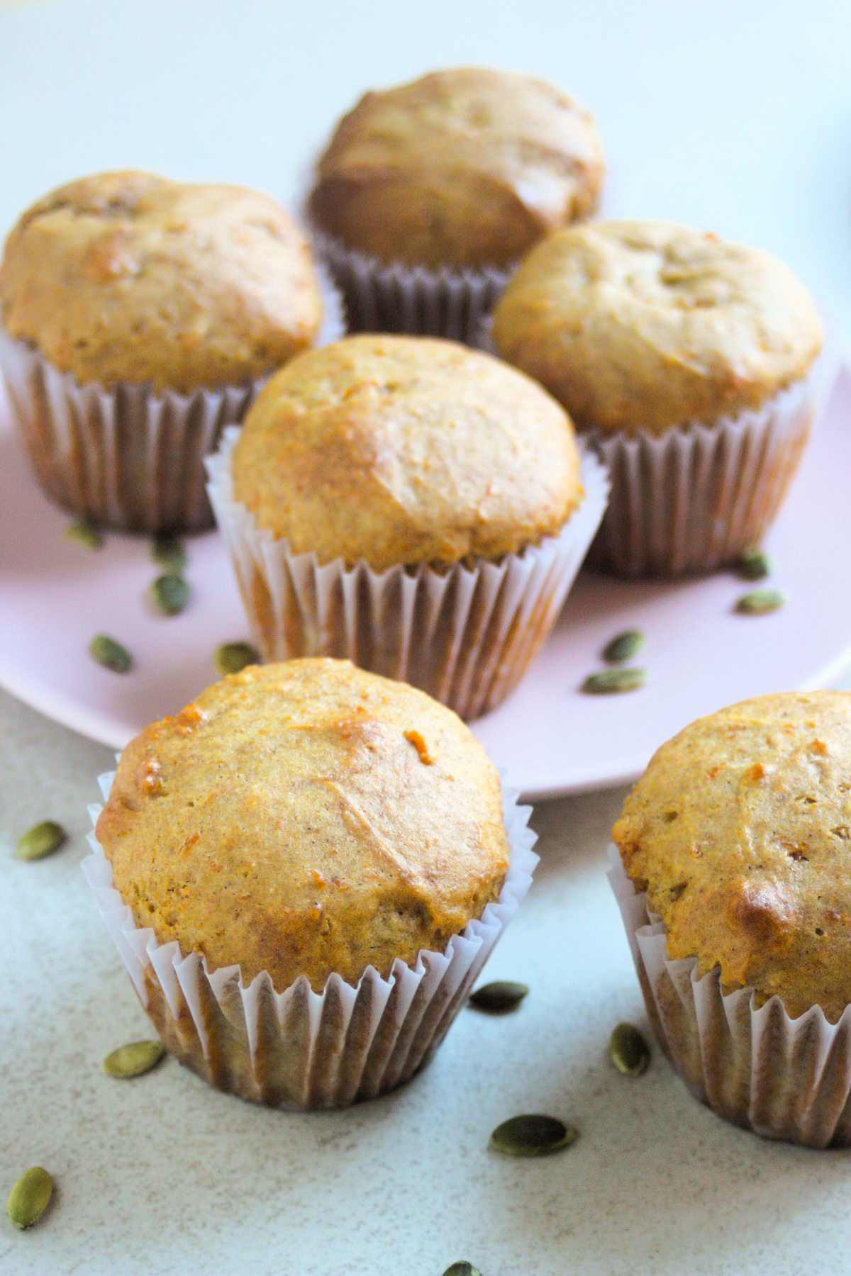 Pumpkin muffins with liners on a white surface, more muffins behind on a pink plate, and pumpkin seeds spread around.