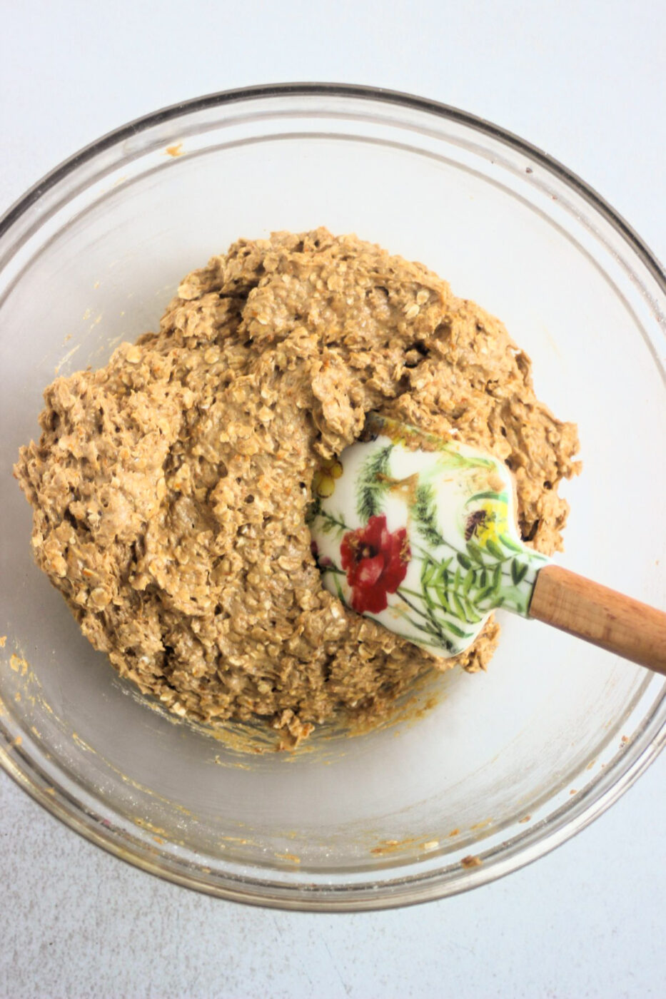 Glass bowl with a light brown mixture, that includes oats, and a rubber spatula.