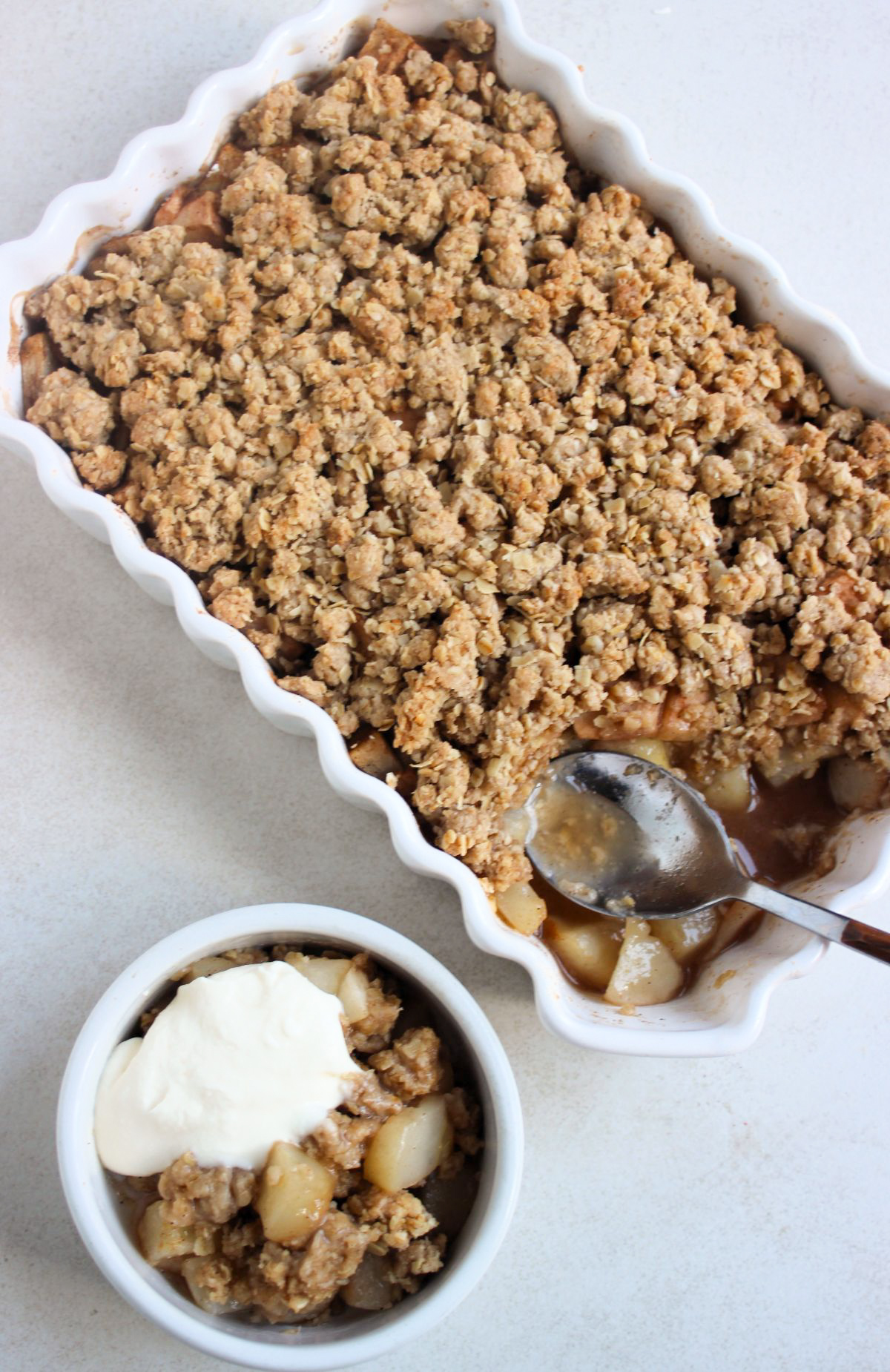 Rectangular baking dish with pear crisp and a spoon seen from above. One part is not complete, and there are chopped apples with juice. Small bowl with pear crisp with whipped cream.