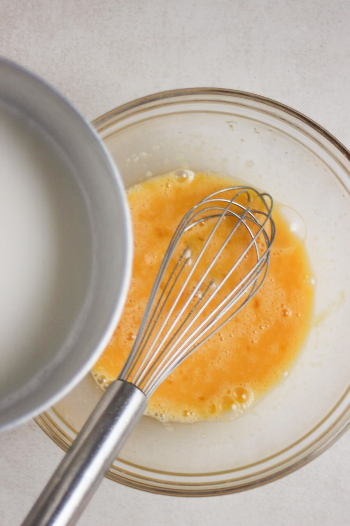 Pouring milk from a saucepan into a bowl with the egg mixture, with a hand whisk nearby.