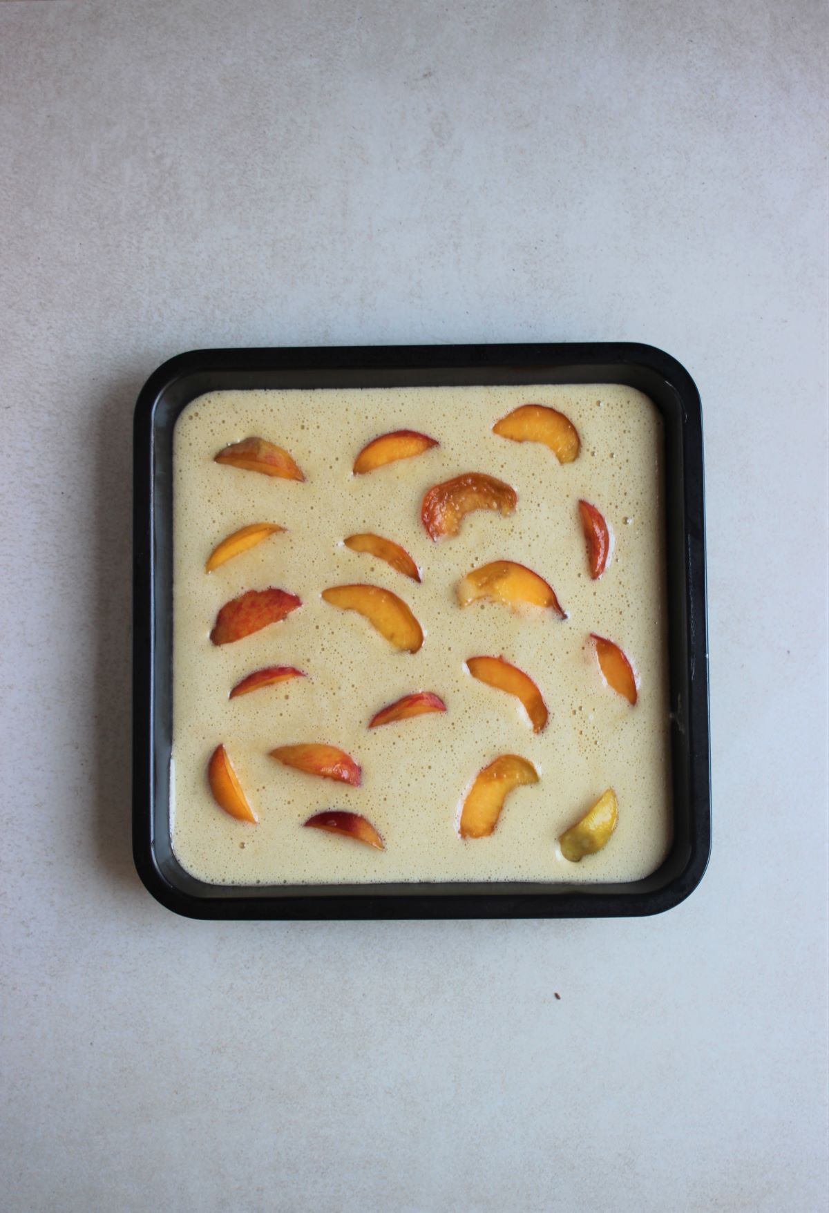 A square bake sheet pan with cake batter and sliced peaches on a white surface.