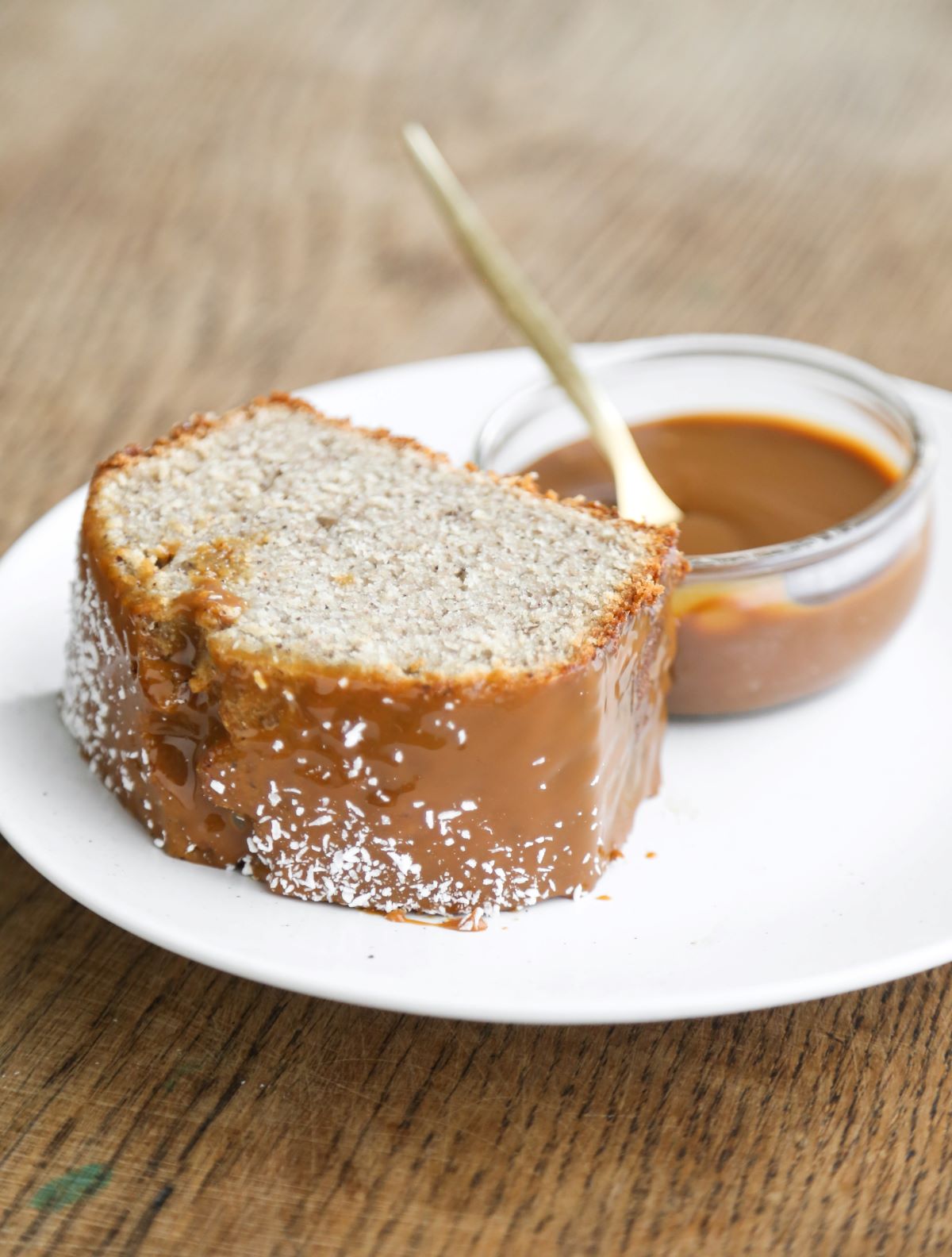 Slice of banana loaf cake with a dip of dulce de leche on a white plate on a wooden surface.