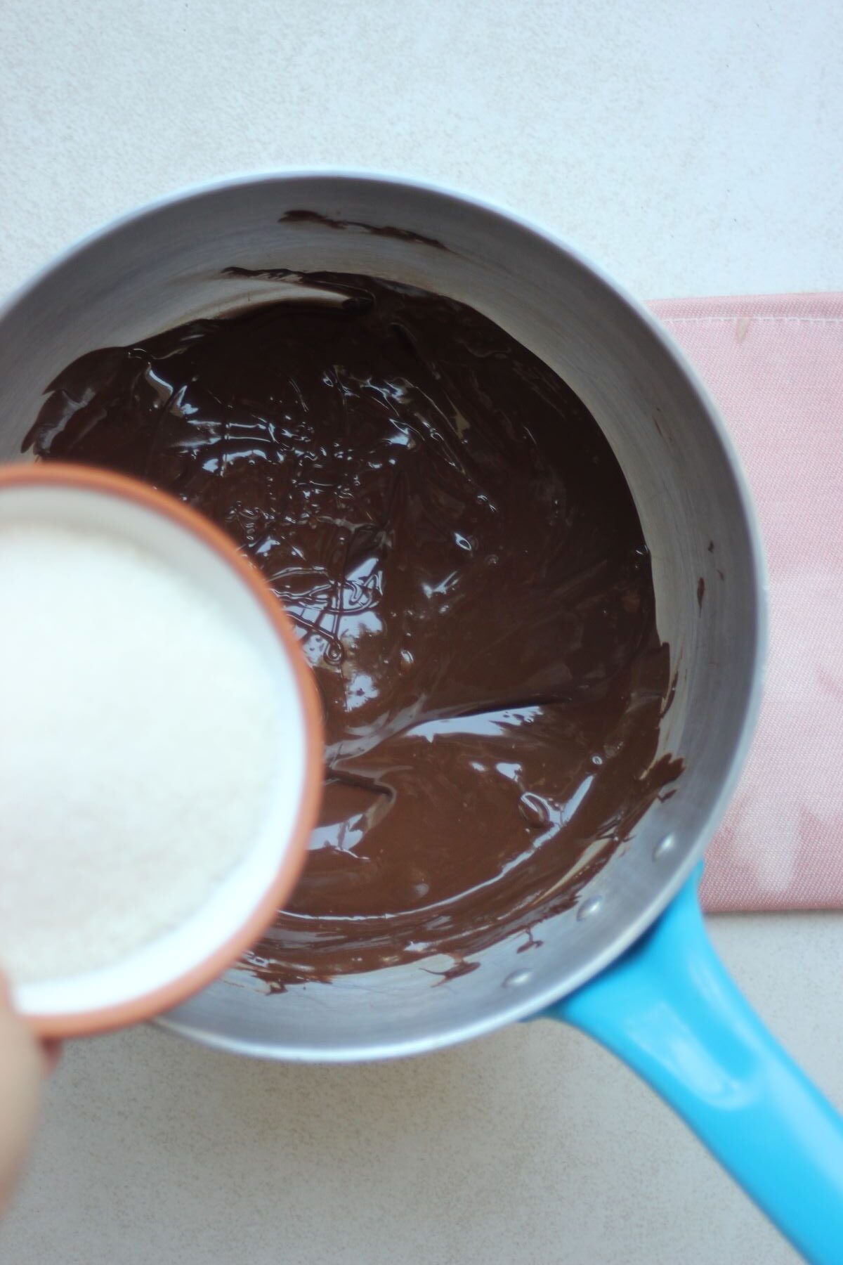 A tiny bowl with sugar is about to be poured into a deep saucepan with melted chocolate.