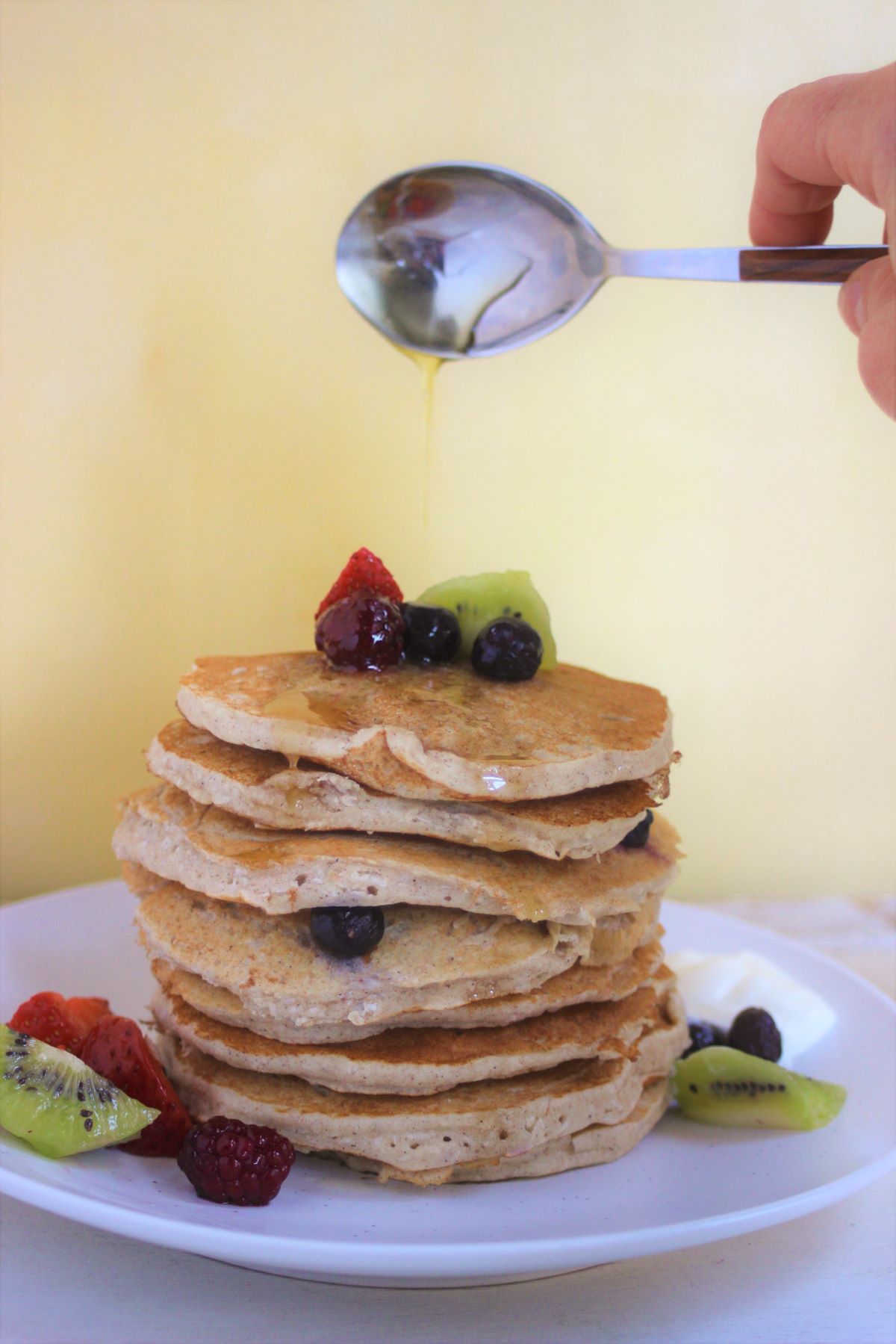 A tower of pancakes with fresh fruits and a spoon throwing honey to it.