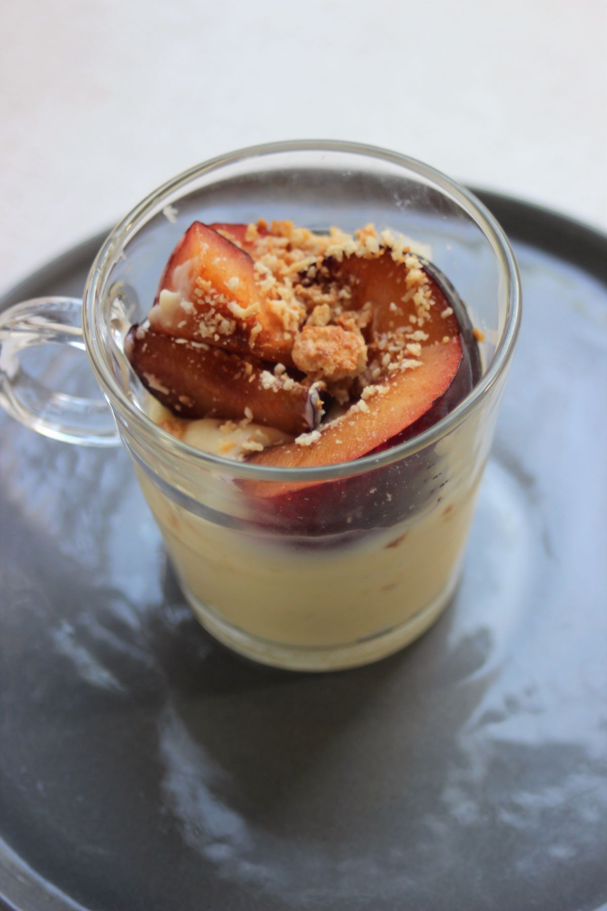 A glass cup with cream (panna cotta), plum slices, and crumbs.