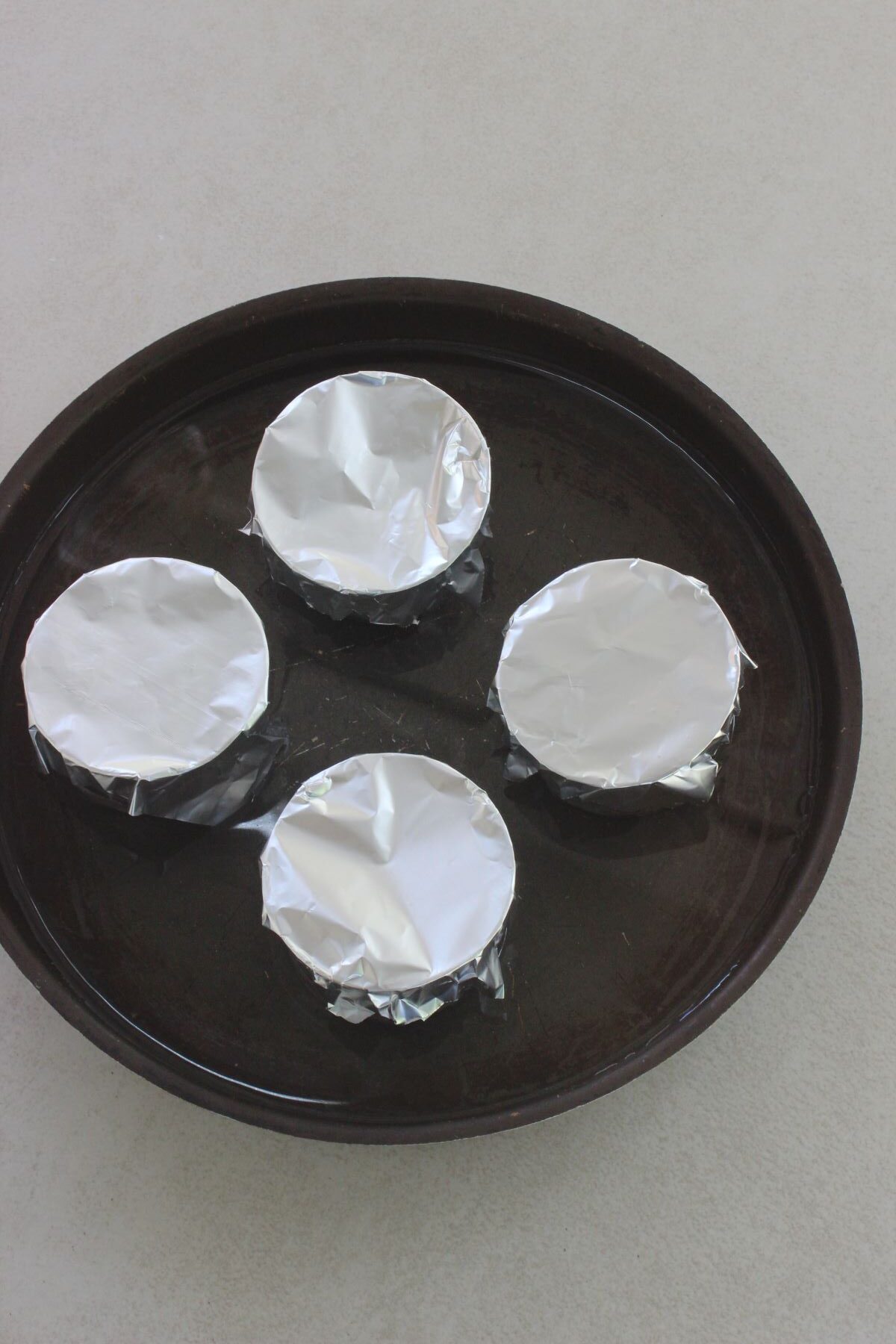 Four flan molds covered with aluminium on a baking sheet with water.