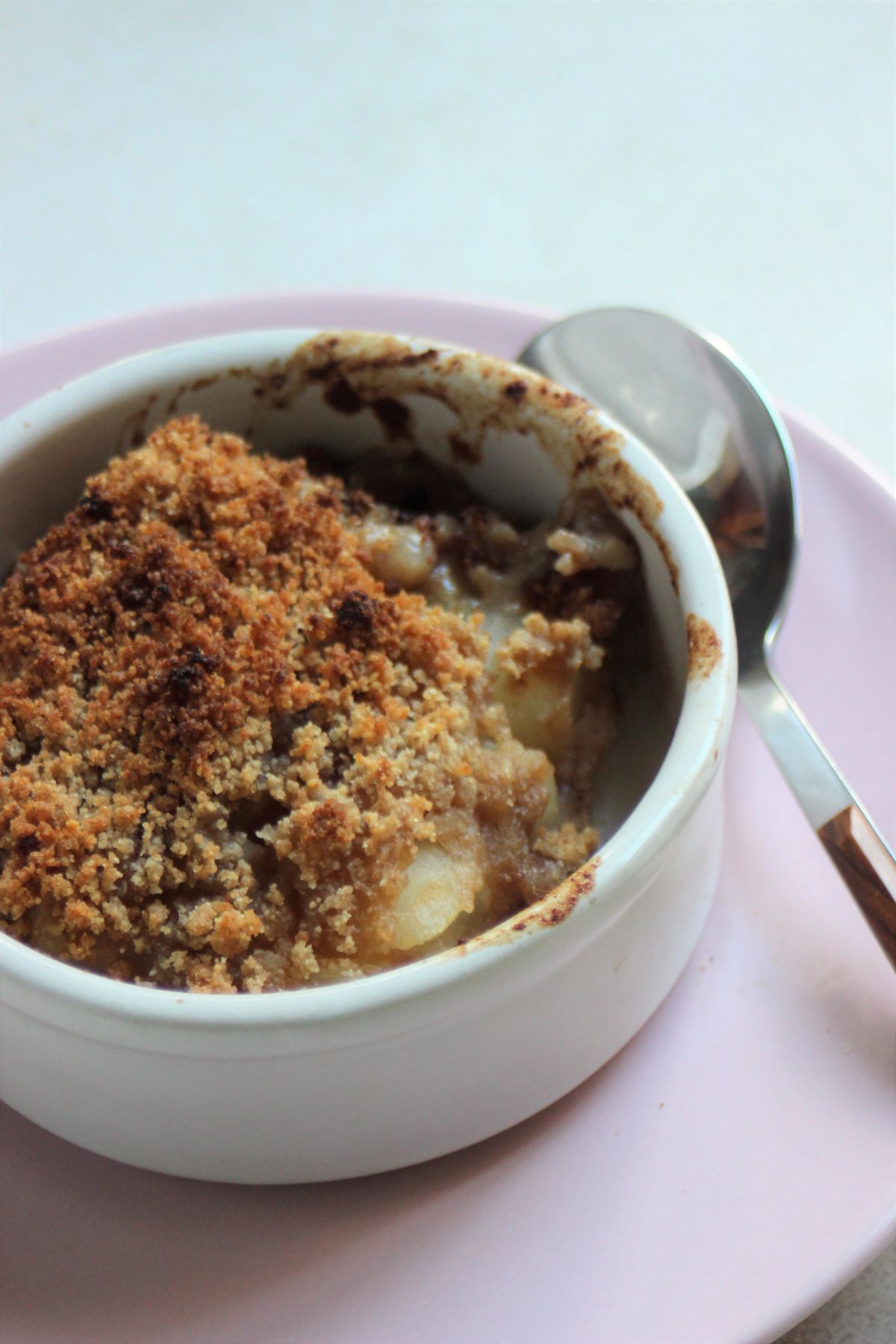 White ramekin with pear crumble and a spoon on the side, on a pink plate.
