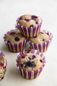 Four mixed berry and lemon muffins with liners on a white surface.