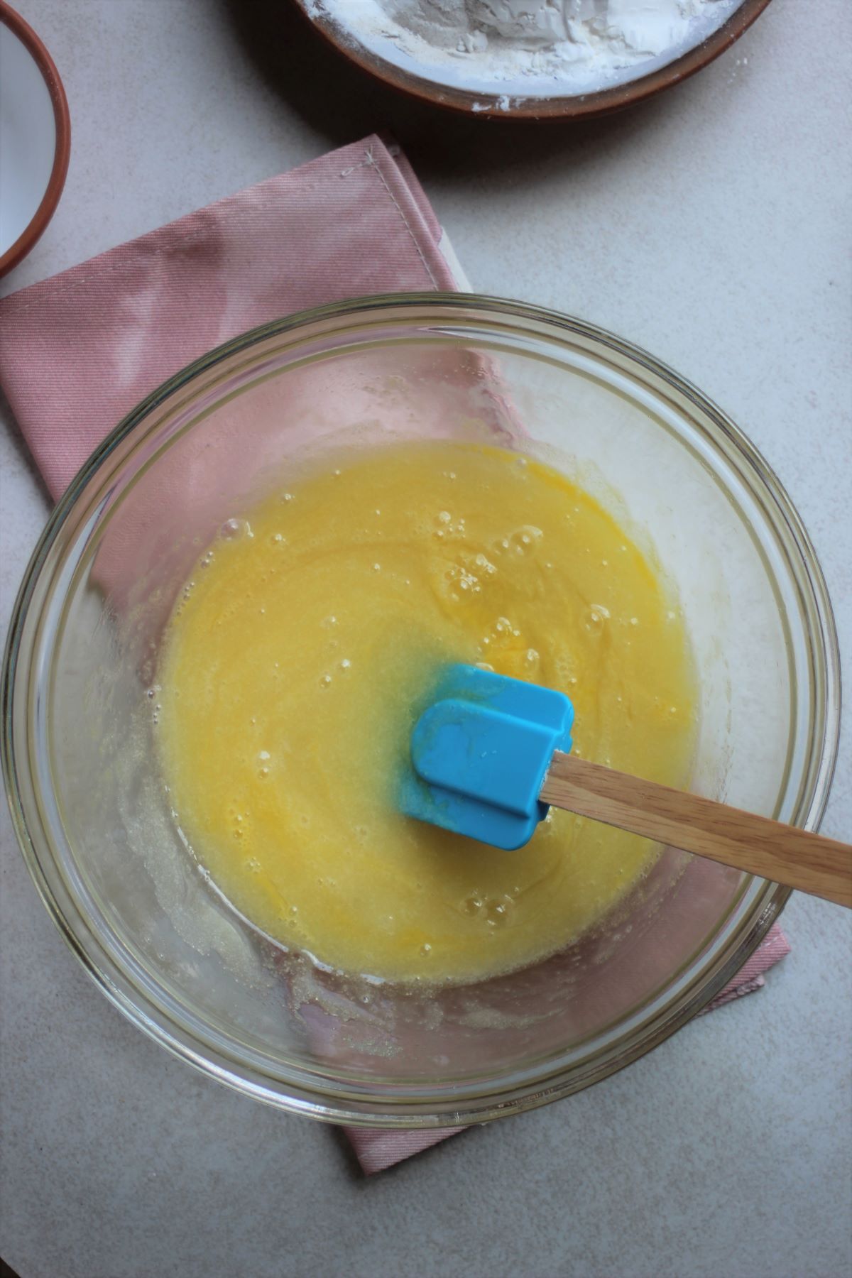 Glass bowl with a yellow mixture and a rubber spatula. Pink napkin under the bowl.