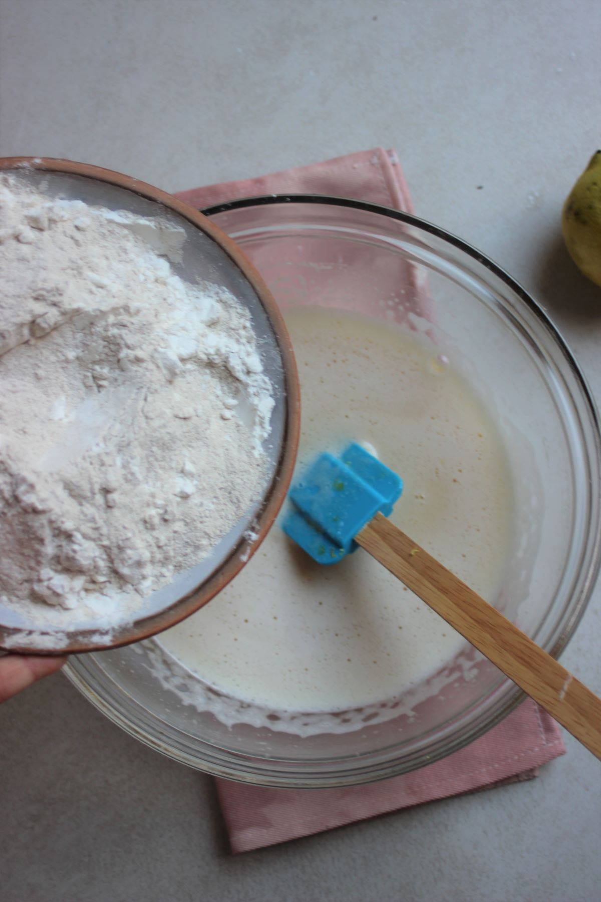 Plate with flour is about to be poured into a glass bowl with a liquid mixture and a rubber spatula.