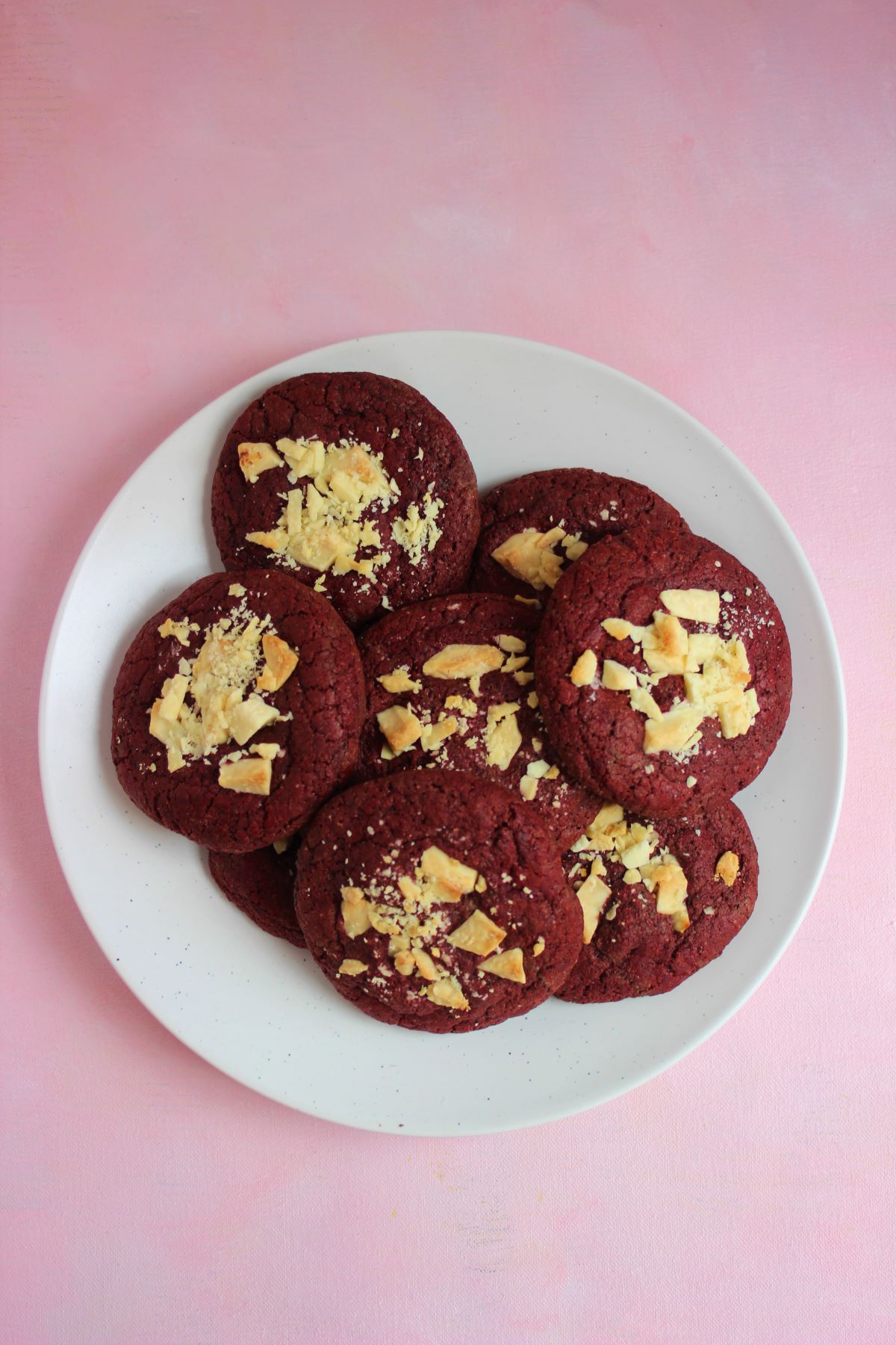 Many red velvet cookies with white chocolate chunks on a white plate seen from above.
