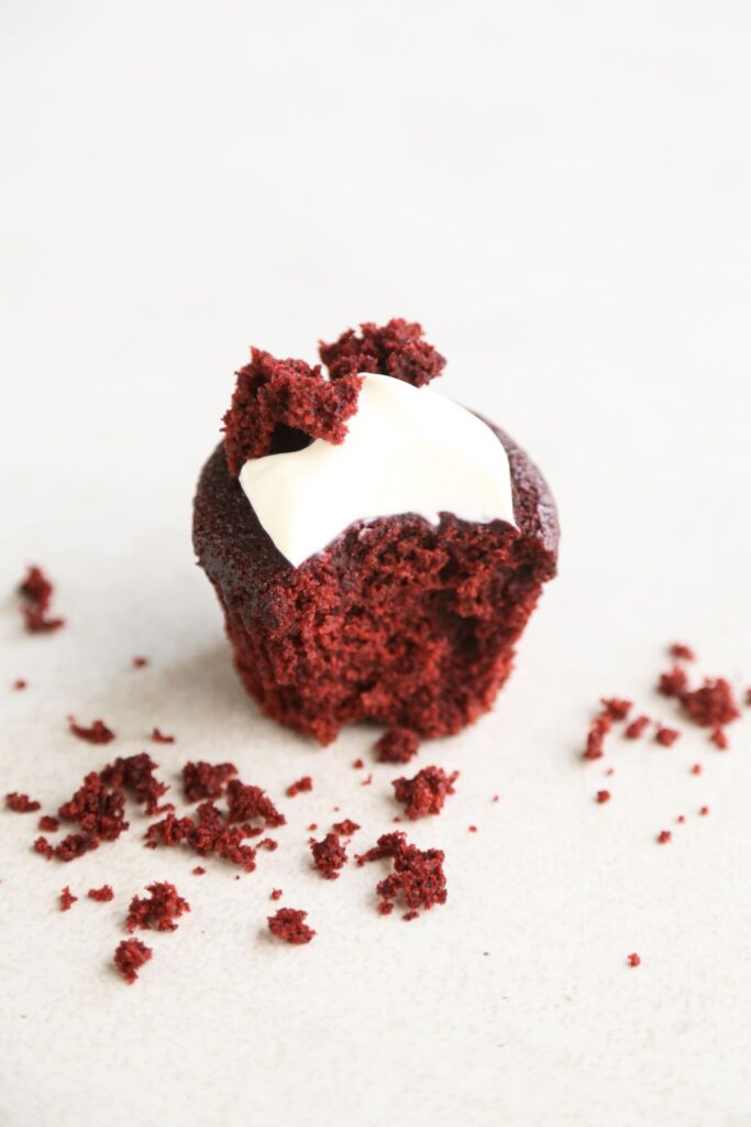 Red velvet muffin with cream cheese frosting without a piece on a white surface.