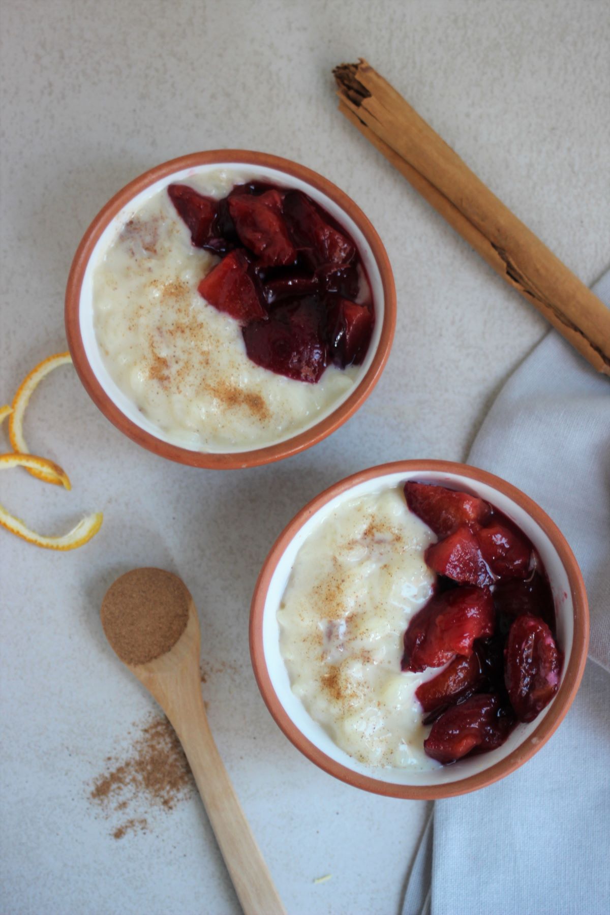 Two tiny bowls with rice pudding and plum compote seen from above. A tablespoon and a cinnamon stick.