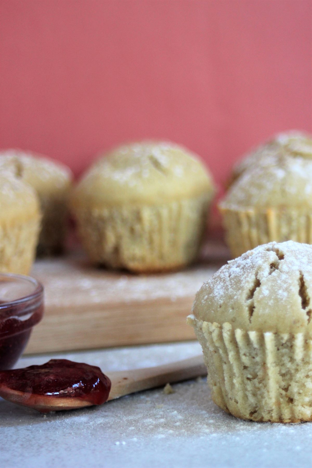 Vanilla muffin and a spoon with red jam. Many muffins behind, on a wooden board.