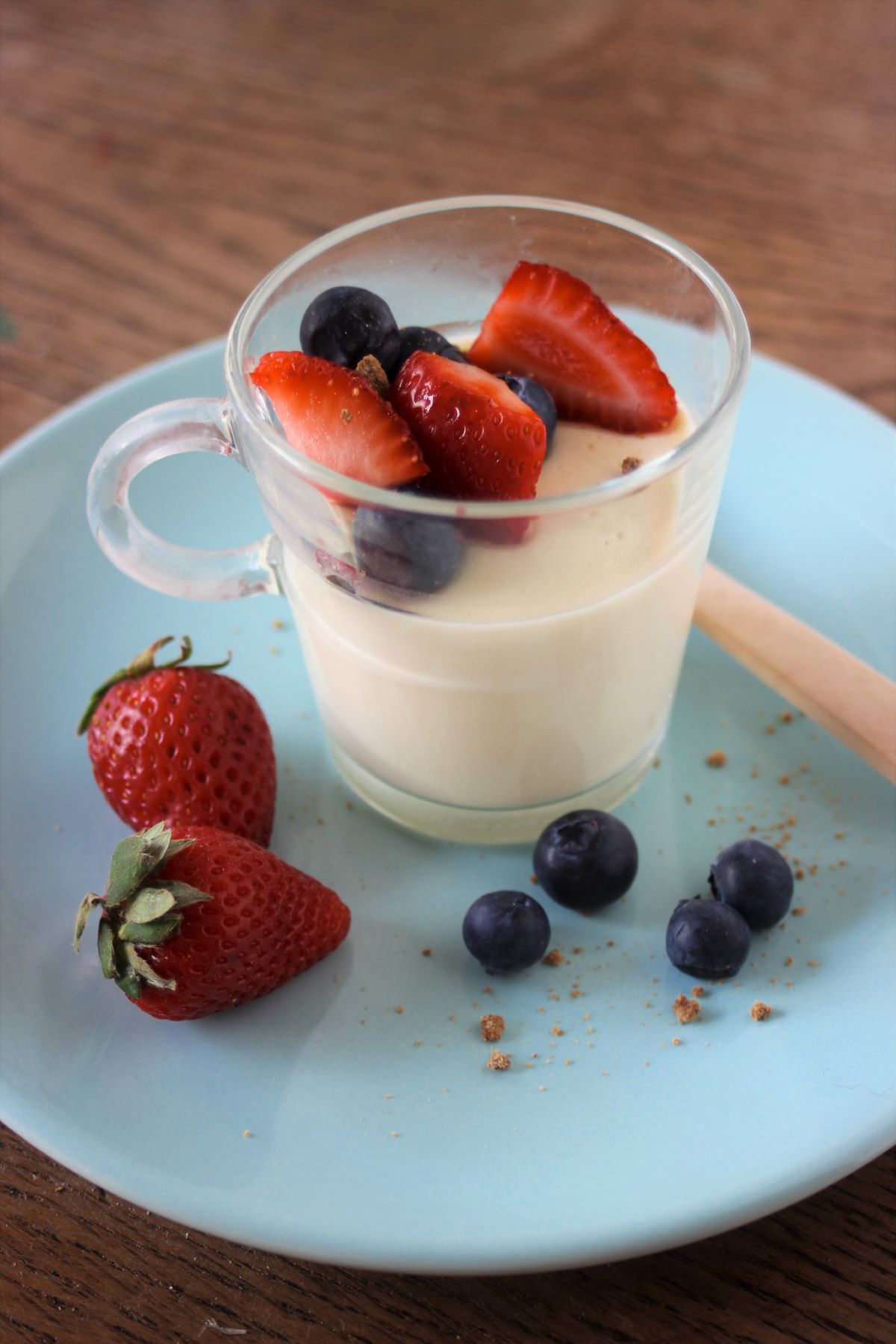Glass cup with vanilla panna cotta, strawberries, and blueberries on a light blue plate.