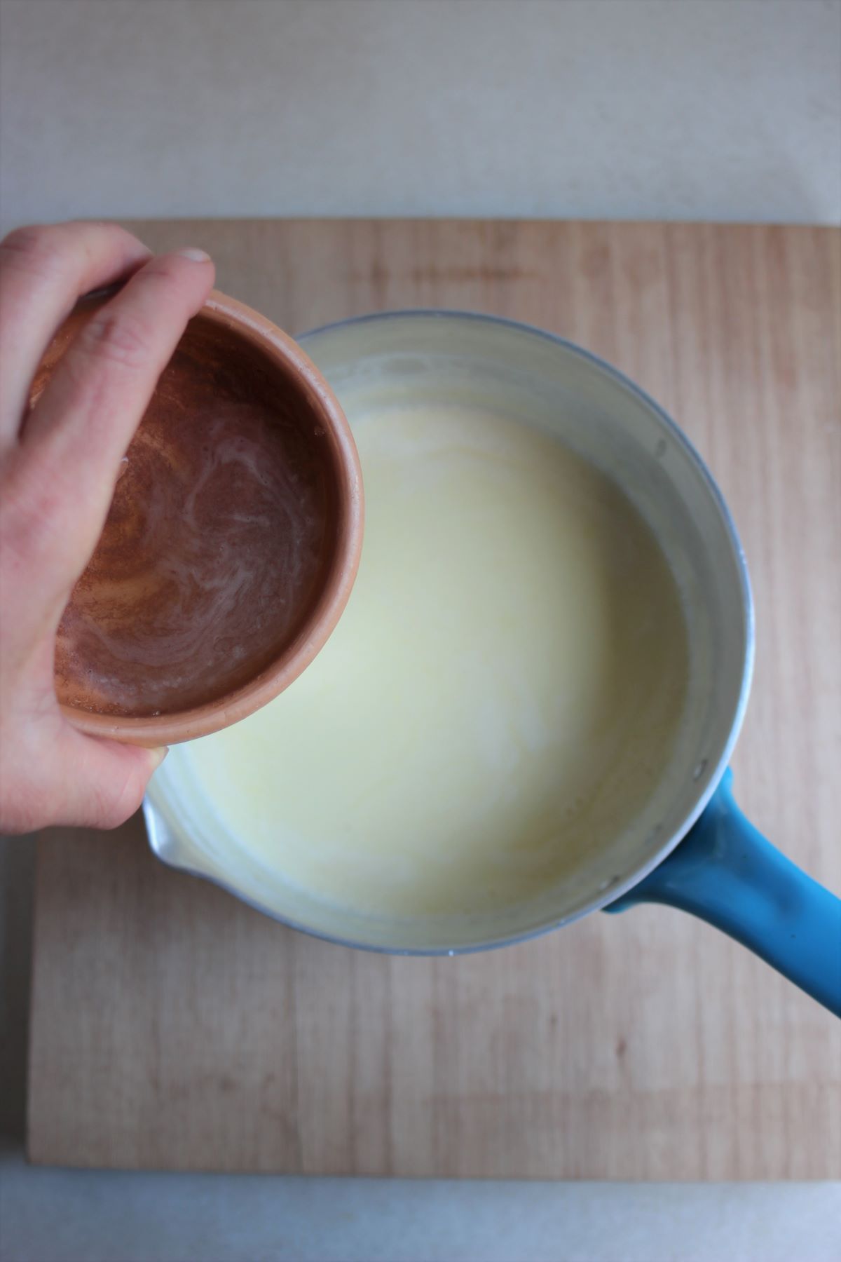 Tiny bowl with a liquid is about to be poured into a deep saucepan with milk.