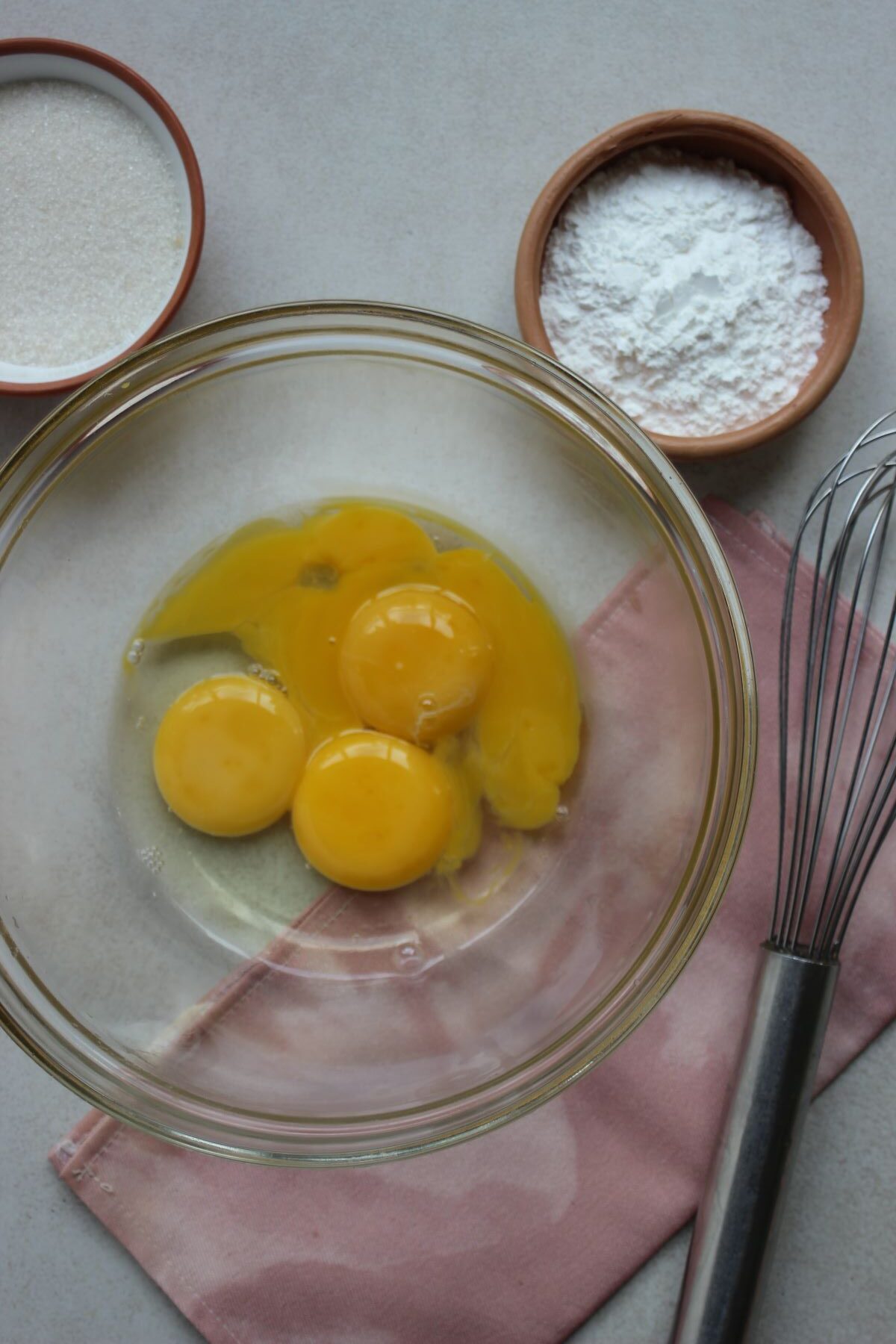 Glass bowl with 3 egg yolks and a hand whisk on the side.