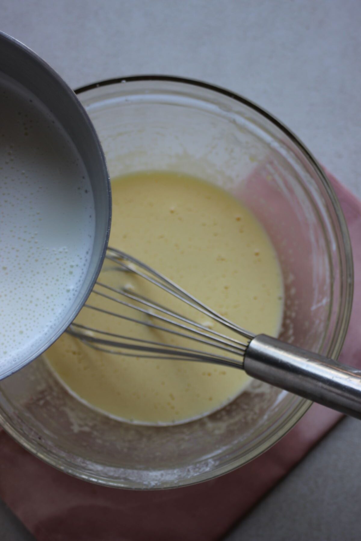 Saucepan with milk is about to be poured into a glass bowl with a liquid and a hand whisk.