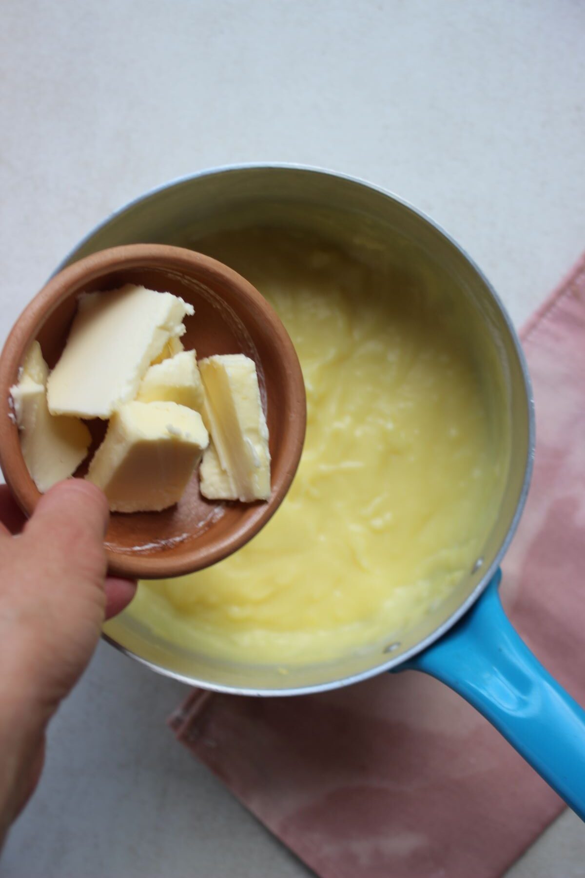 A tiny bowl with butter is about to be poured into a deep saucepan with pastry cream.