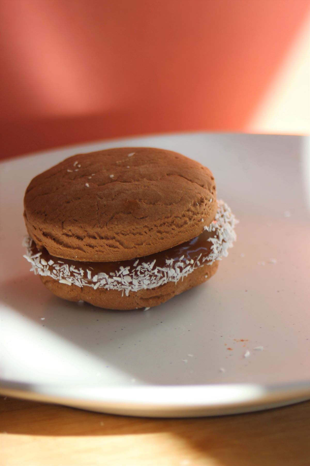 A chocolate alfajor with dulce de leche and shredded coconut on a white plate.