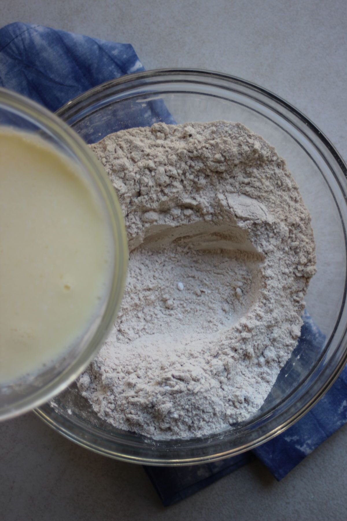 Bowl with white liquid is about to be poured into a glass bowl with flour.