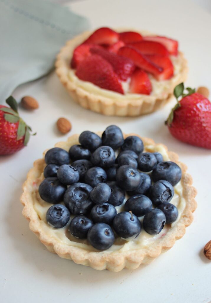 A blueberry tart, and a strawberry tart behind. Fresh strawberries on the sides.