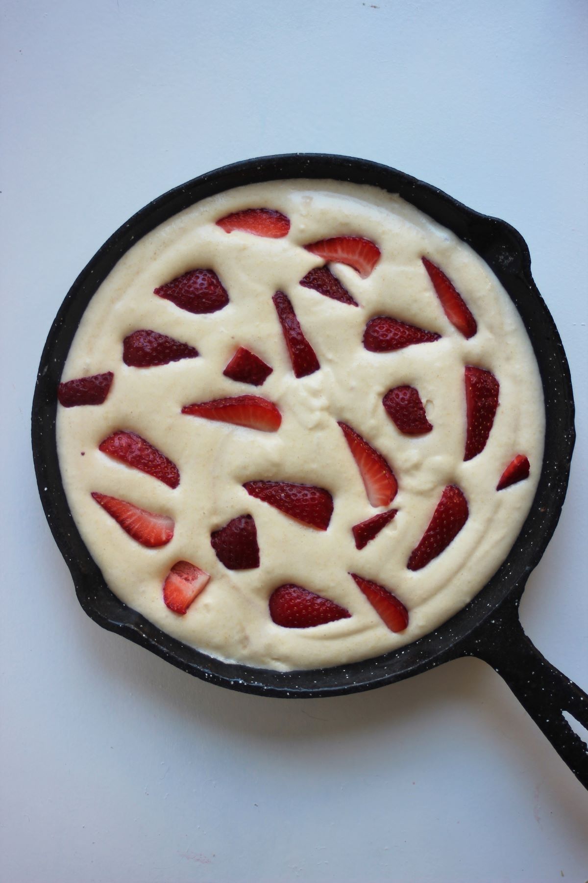 A black skillet with raw strawberry clafoutis on a white surface.