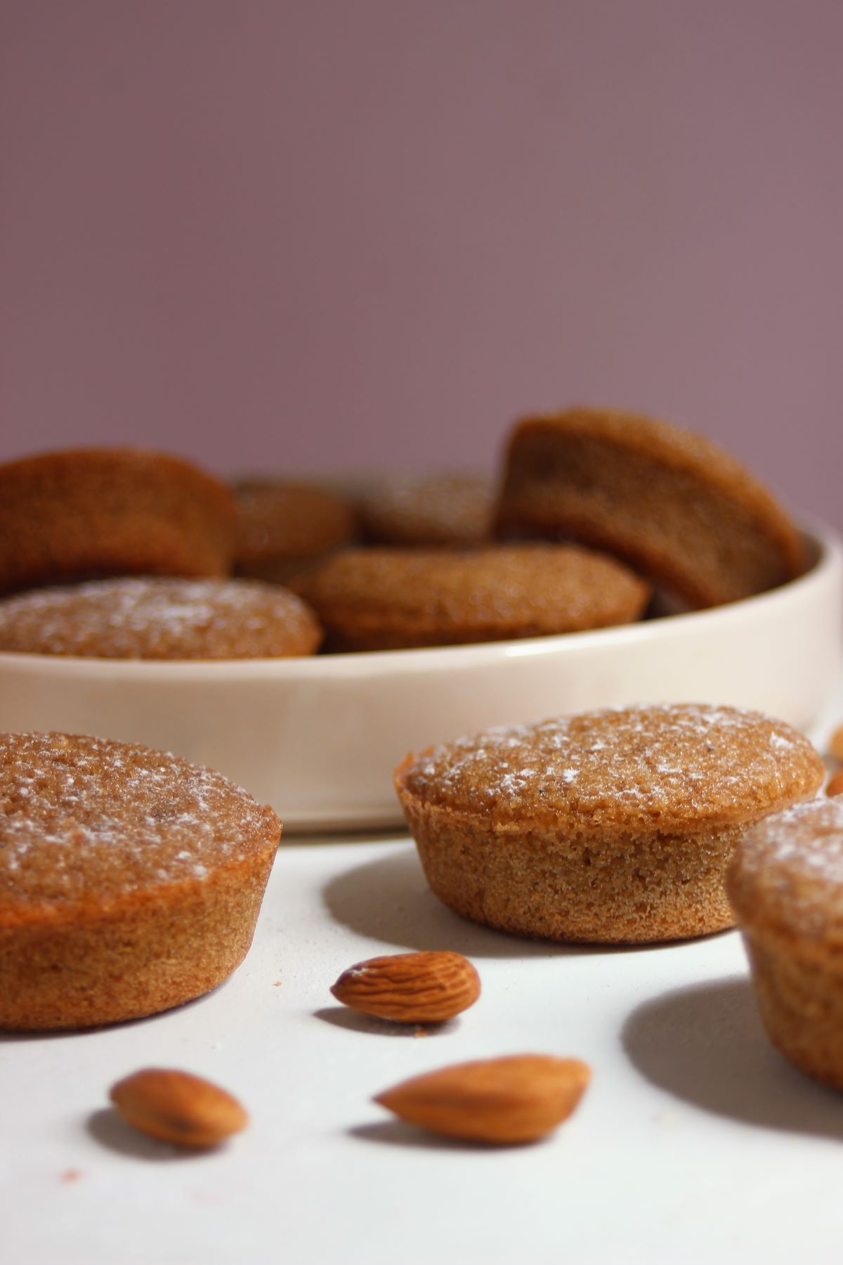 Financiers and almonds on a white surface. Many financiers are in the background on a plate.
