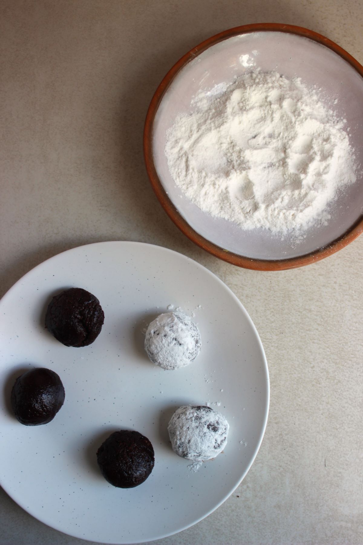 Plate with cookie dough balls, two covered with sugar powdered. Another plate with powdered sugar.