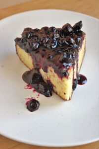 A piece of ricotta cake with blueberry sauce on a white plate.