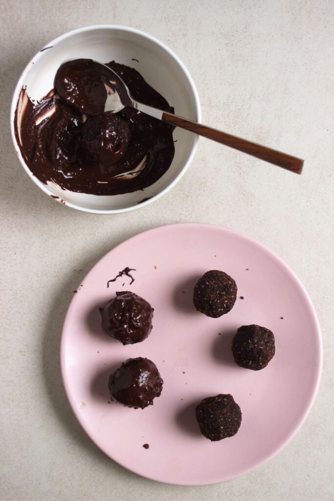 Chocolate peanut butter balls on a pink plate. Another bowl with melted chocolate and a spoon.